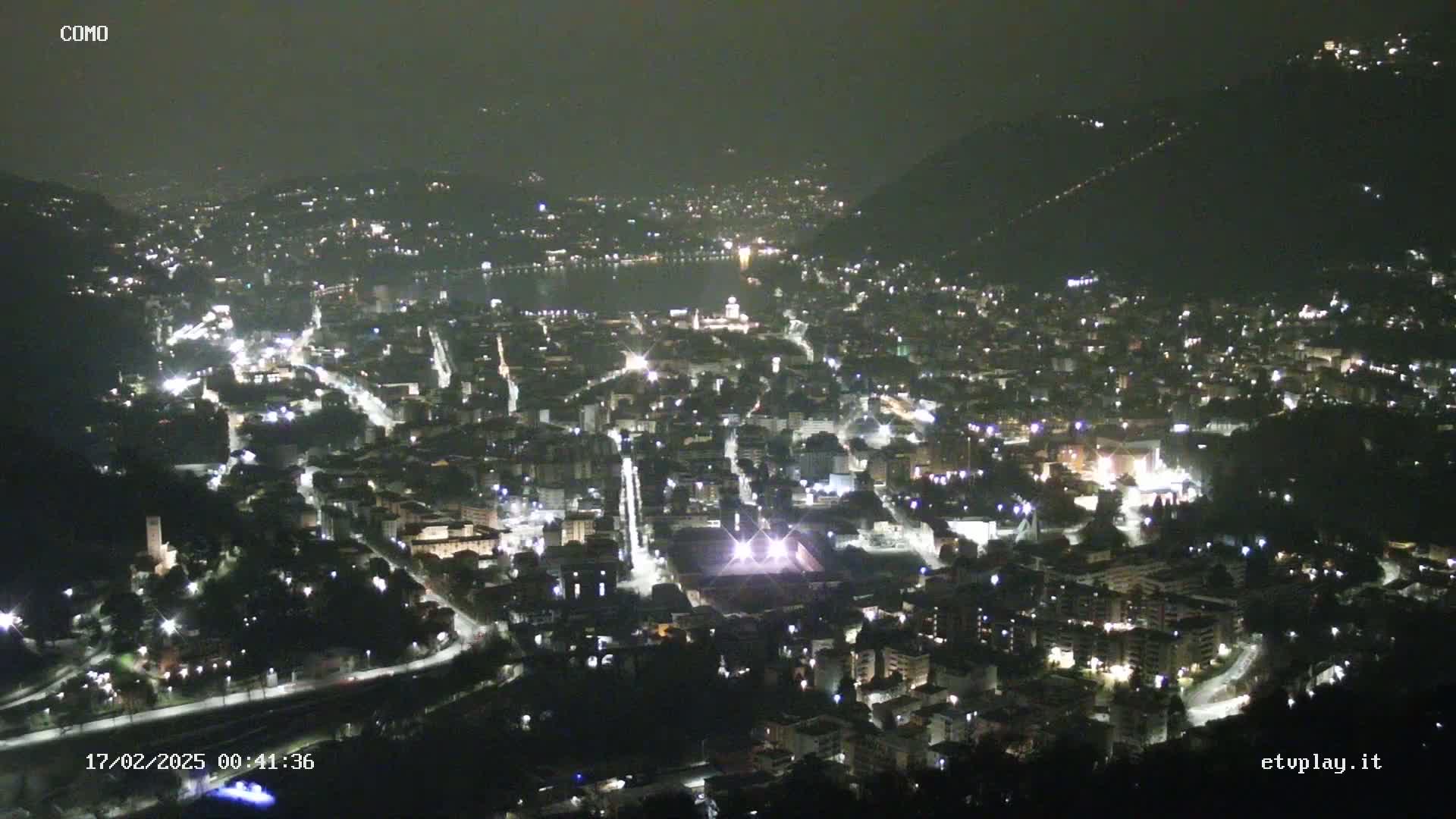 A nighttime aerial view of a city nestled in a valley, with numerous lights illuminating buildings and streets.