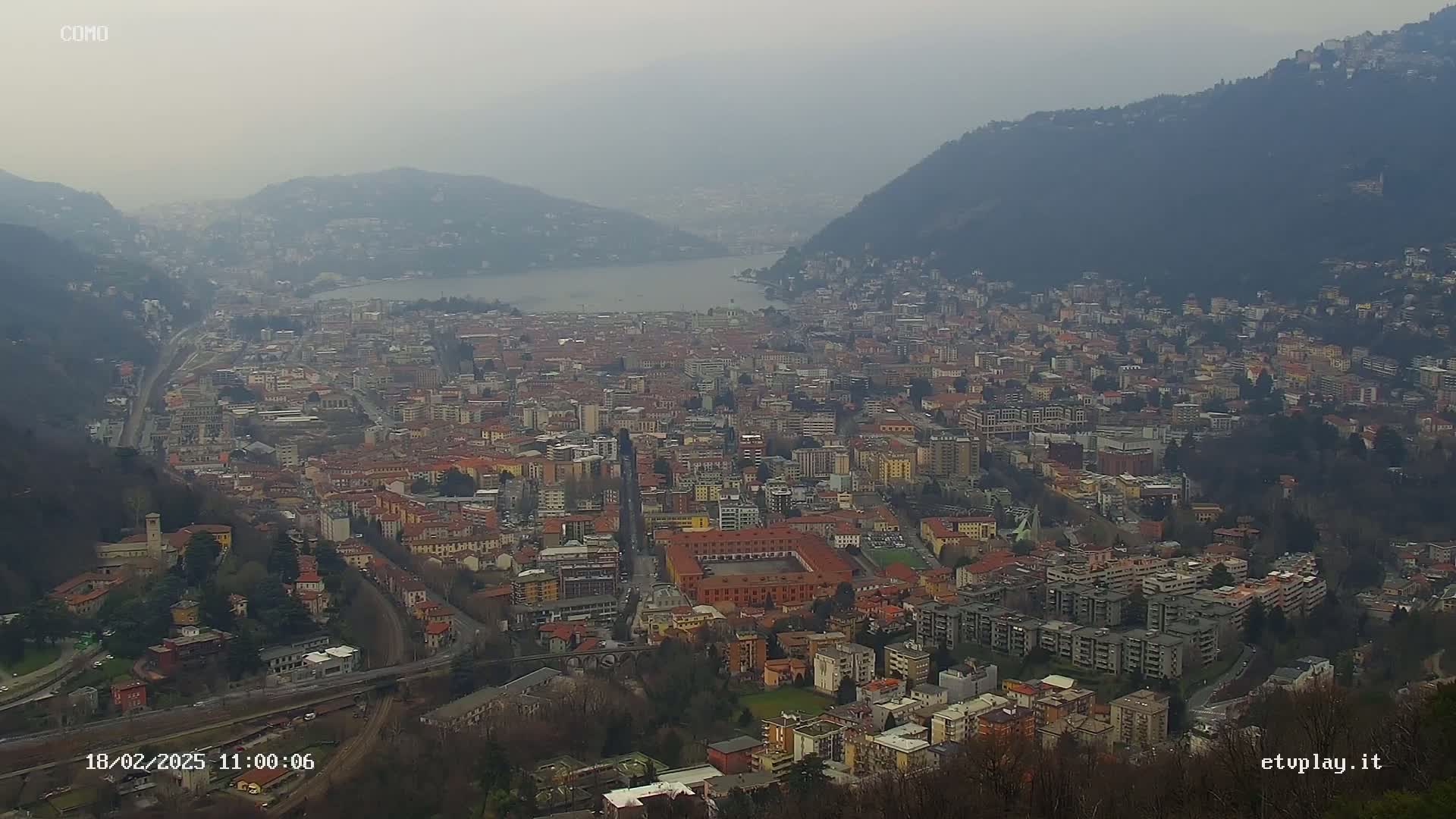 A hazy, high-angle view shows a sprawling city nestled between hills and a lake on a cloudy day.