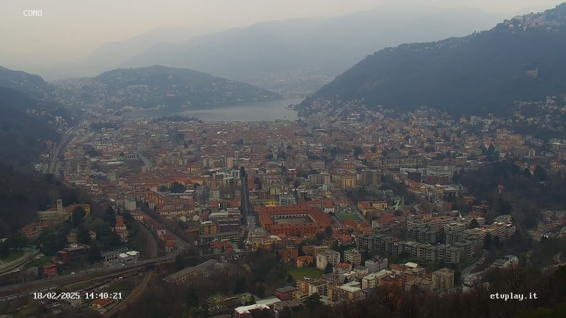 A hazy, overcast view shows a large city nestled between mountains and a lake.