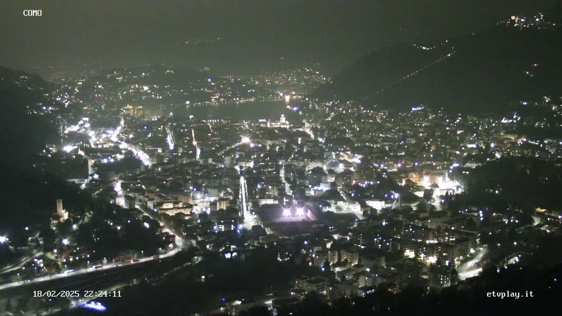 A nighttime, high-angle view of a city illuminated by lights, nestled between hills and near a body of water.