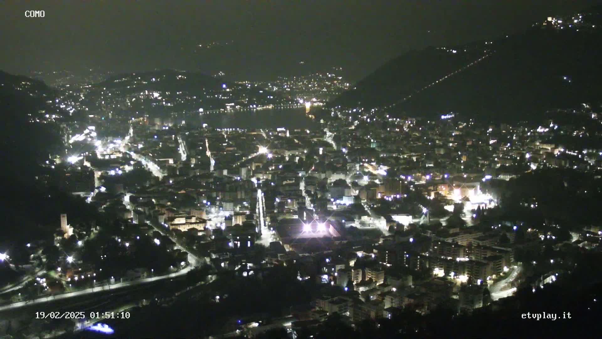 A nighttime, aerial view of a city nestled in a valley, illuminated by numerous streetlights, with a body of water visible in the distance.