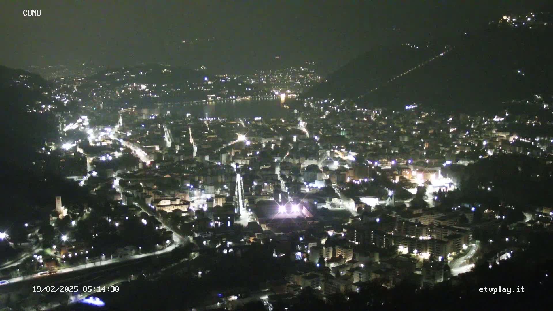 A nighttime aerial view of a city nestled between hills, illuminated by streetlights, with a body of water visible in the distance.