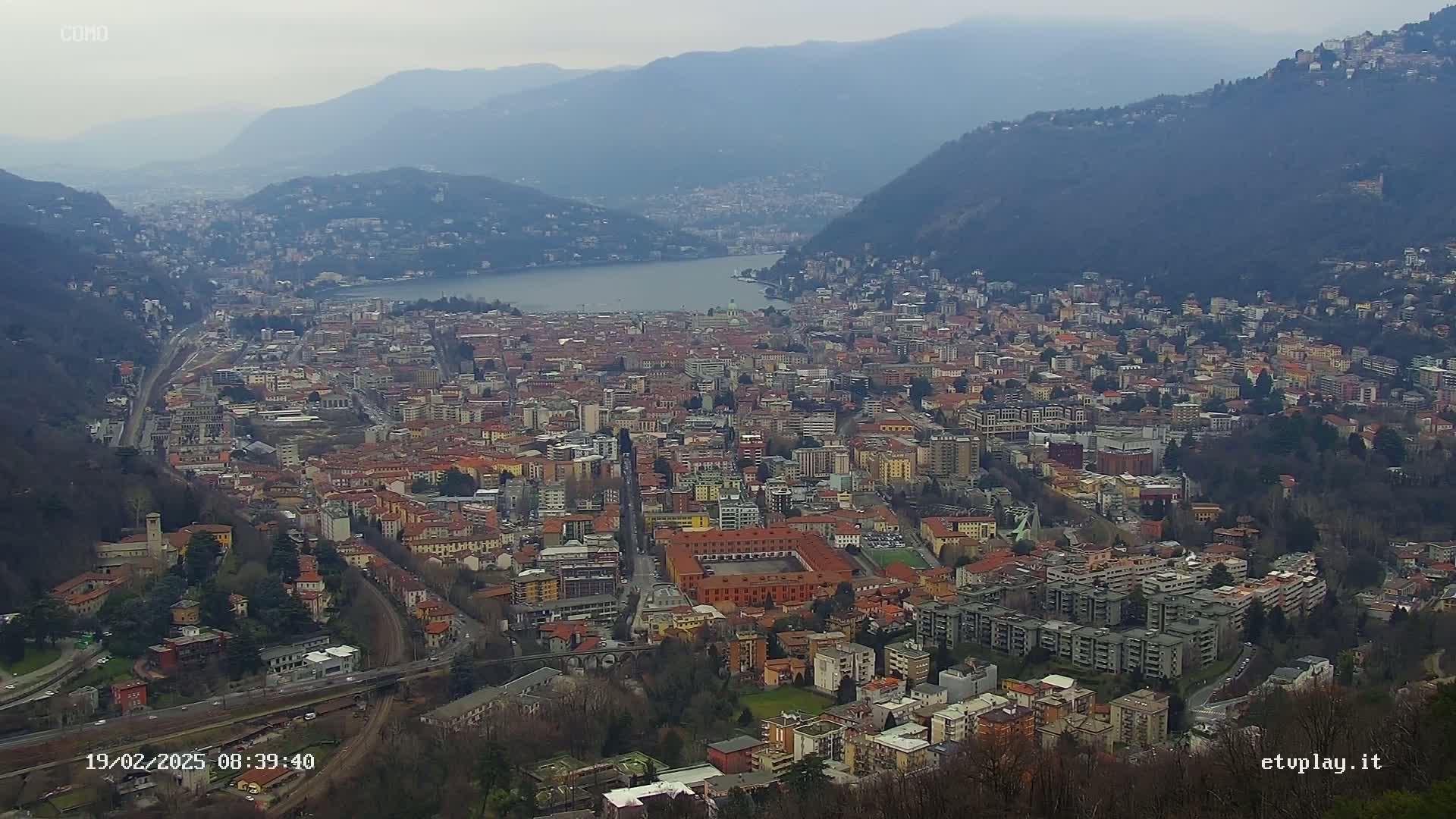 A sprawling city nestled between mountains and a lake under an overcast sky.