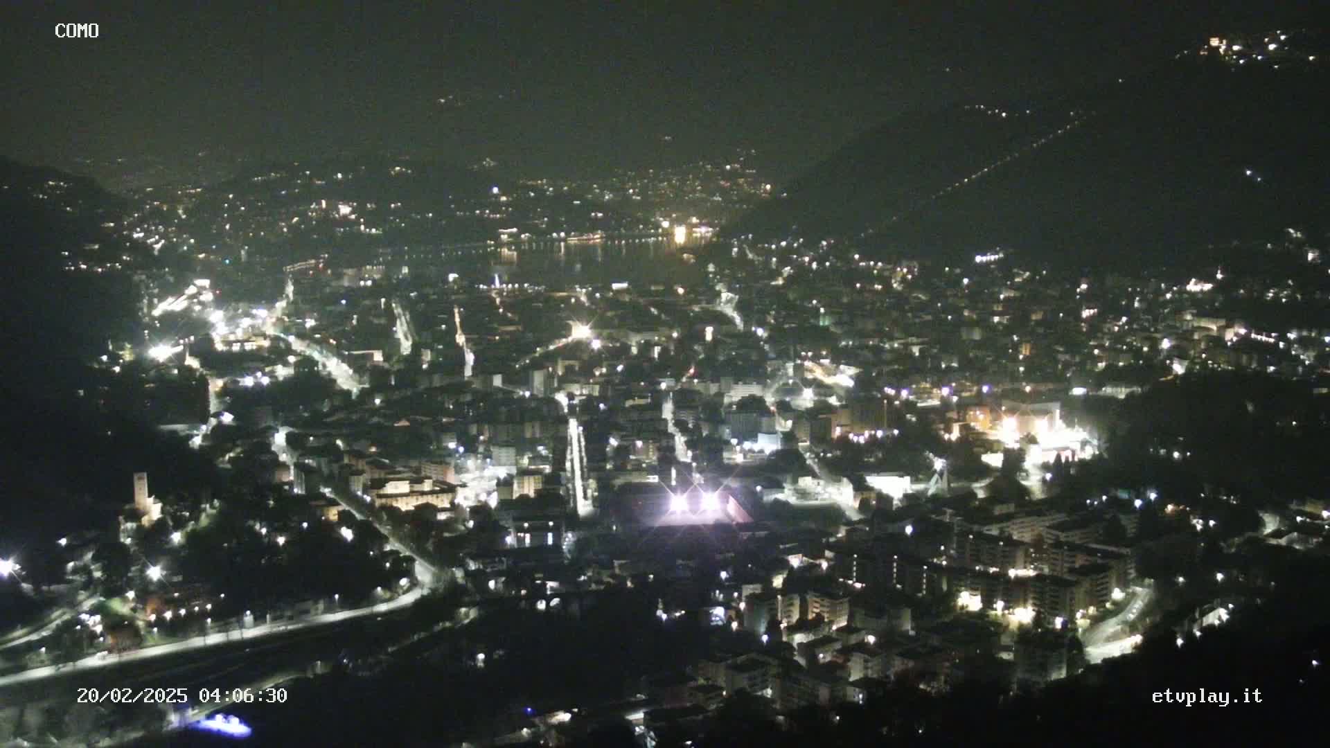 A nighttime aerial view of a city illuminated by streetlights, nestled in a valley beside a body of water under a clear, dark sky.
