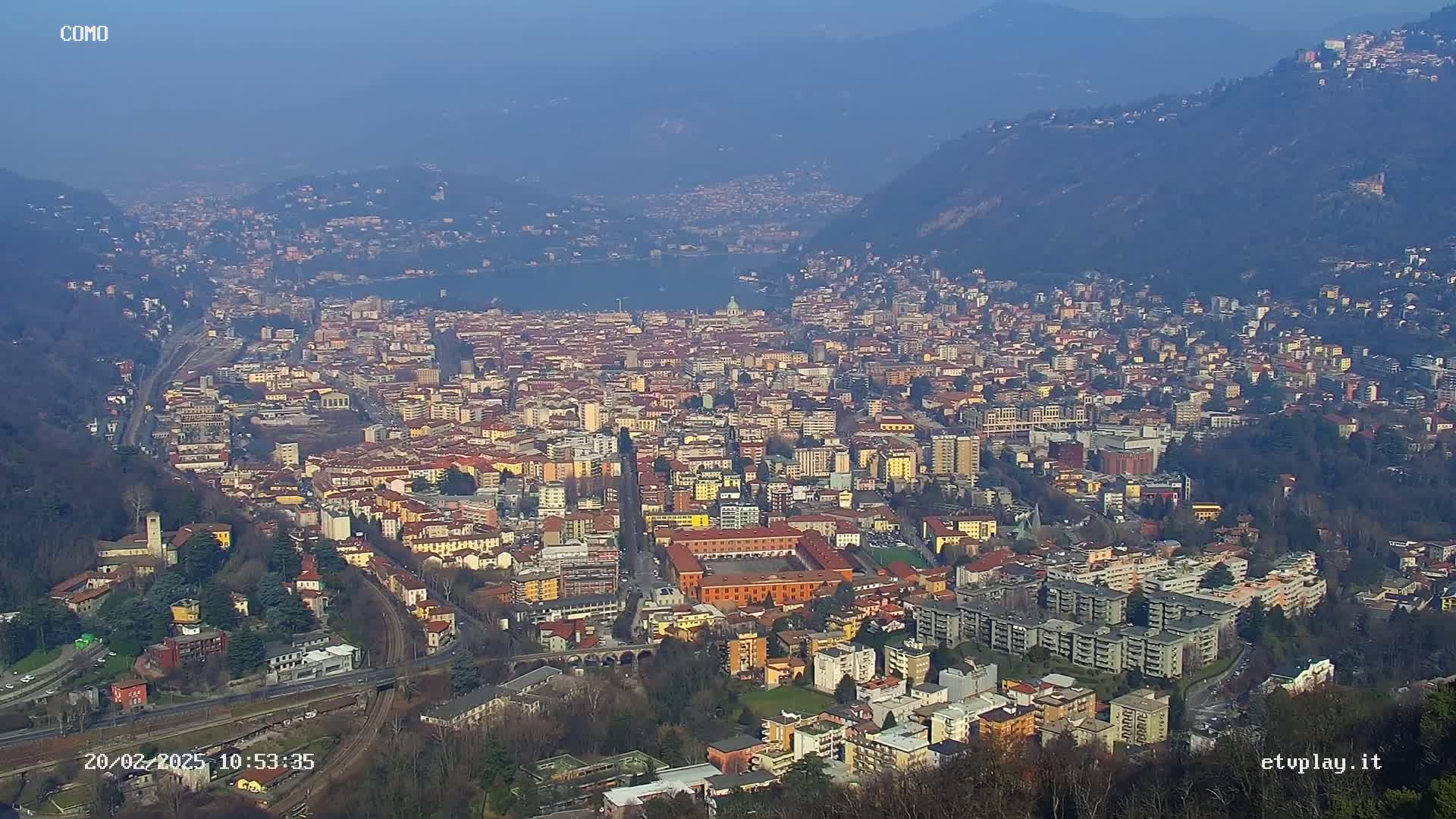 A sprawling city with numerous buildings of varying heights, nestled between hills and a lake under a hazy, clear sky.