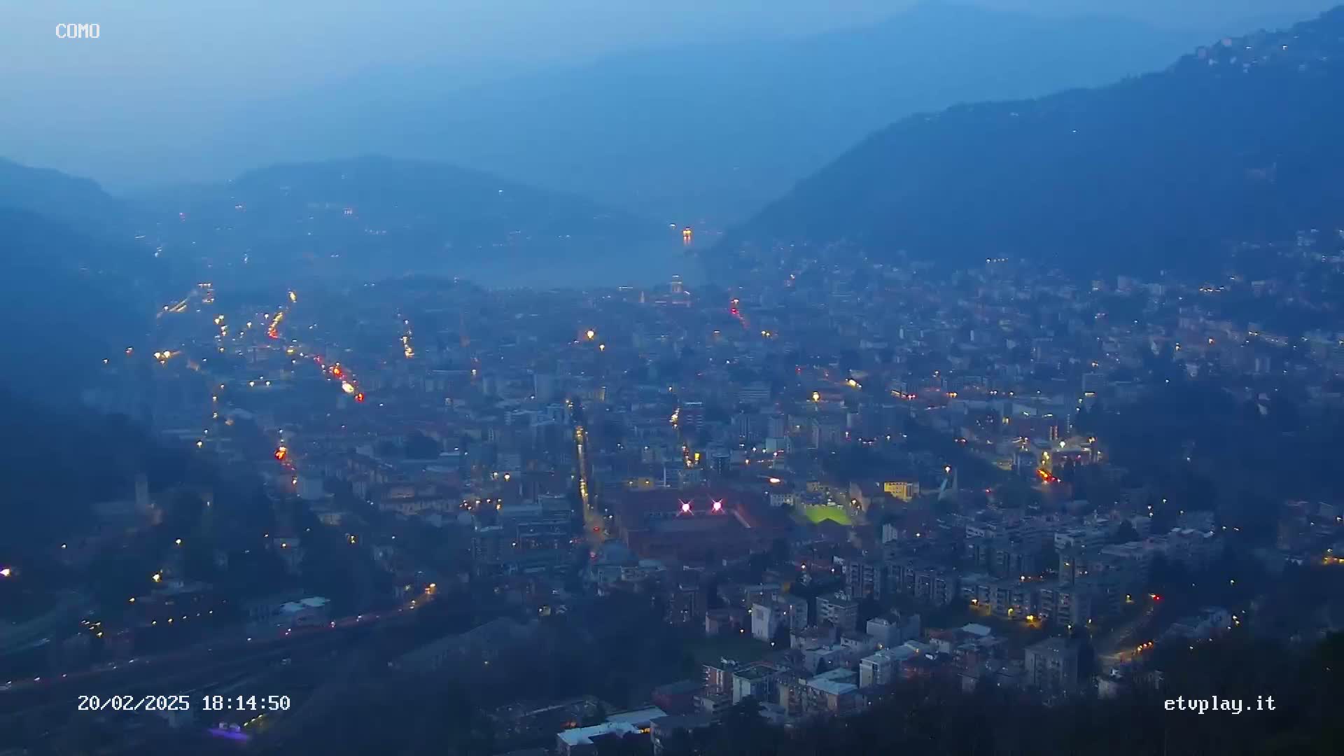 A nighttime, hazy view of a city nestled in a valley between mountains, with scattered lights illuminating the cityscape.