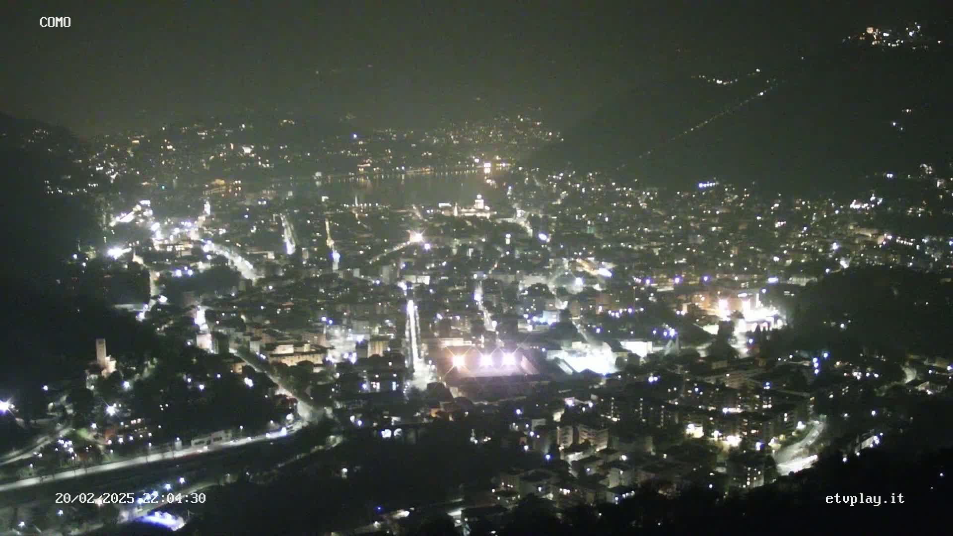 A nighttime, high-angle view of a city illuminated by streetlights and other sources, situated on a hillside next to a body of water.