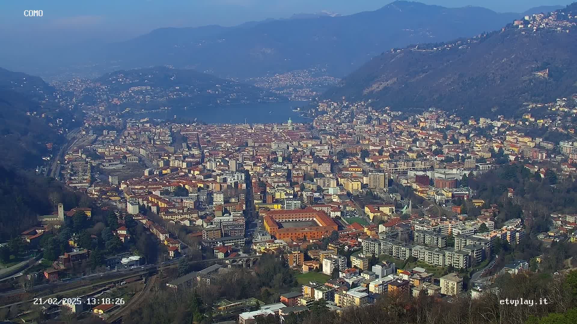 A sprawling city nestled in a valley between mountains, overlooking a lake under a clear, sunny sky.