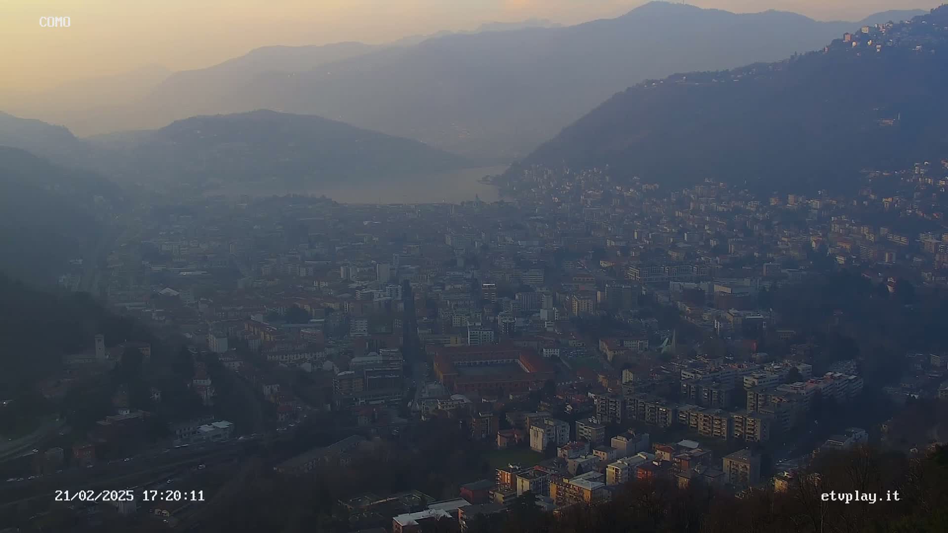 A hazy, dusky view overlooks a sprawling city nestled between mountains and a lake.