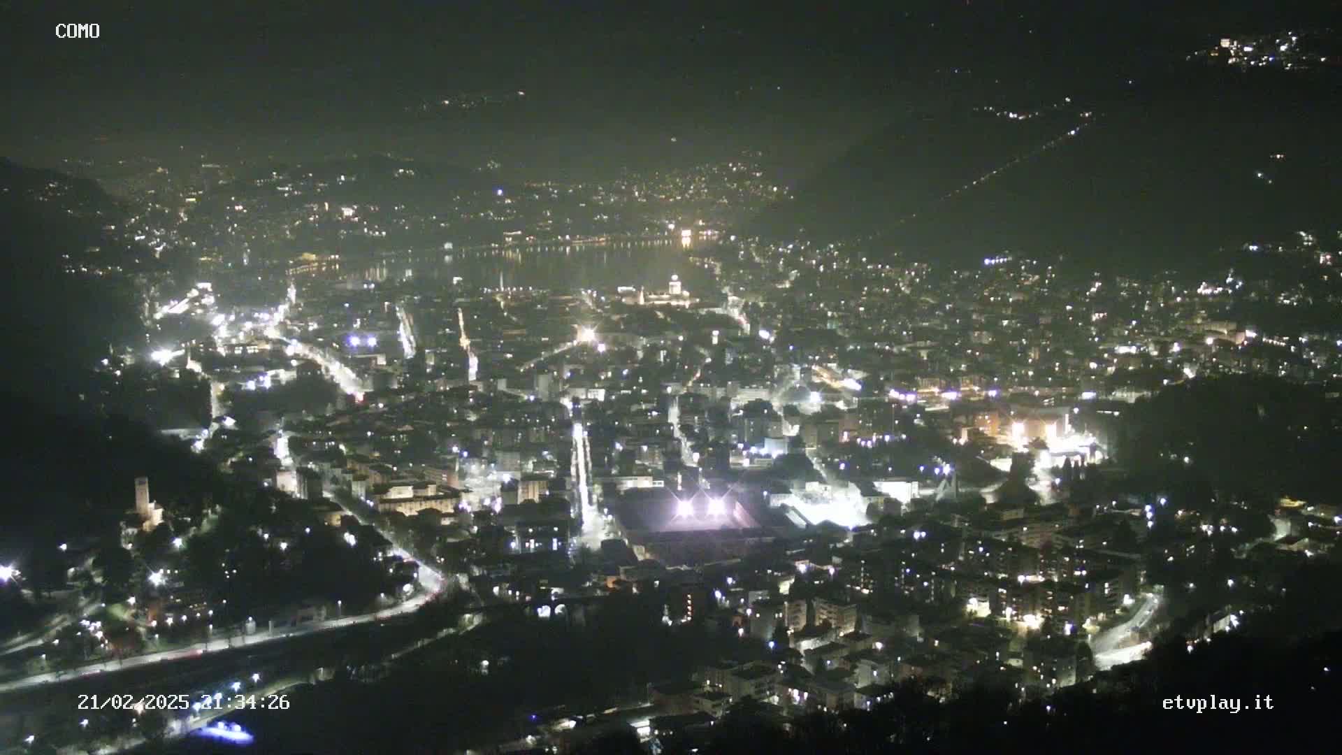 A nighttime aerial view of a city illuminated by lights, situated near a body of water under a clear, dark sky.