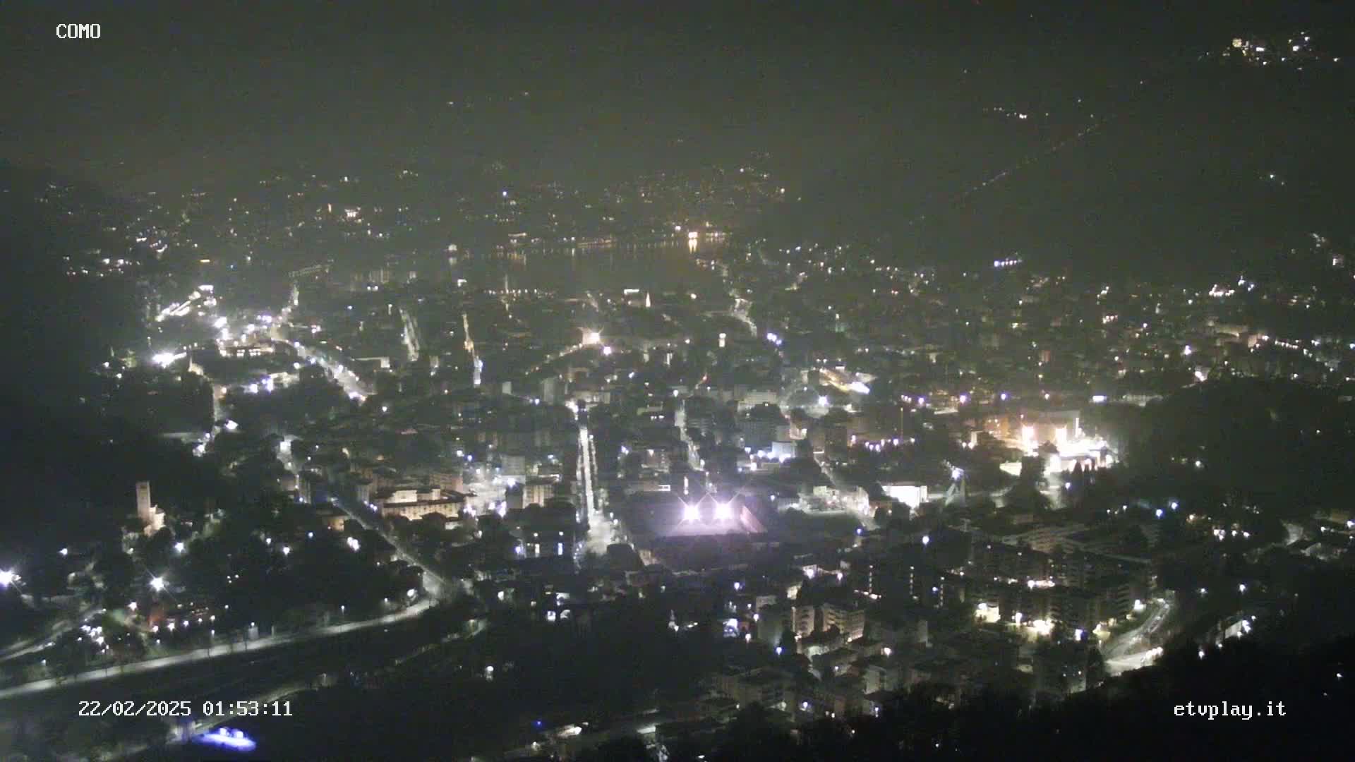 A nighttime, high-angle view of a city illuminated by streetlights, with a body of water visible in the distance.