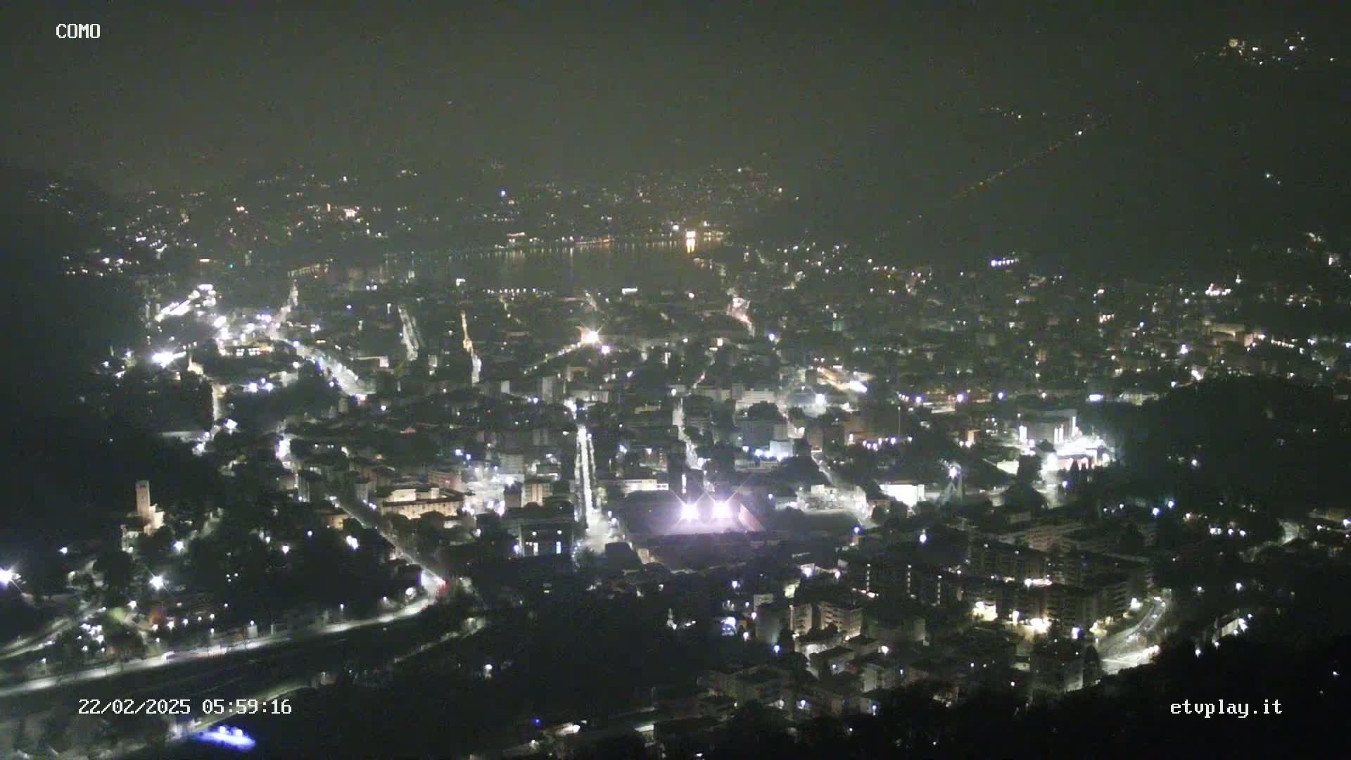 A nighttime, high-angle view of a city illuminated by streetlights, situated near a body of water under a hazy sky.