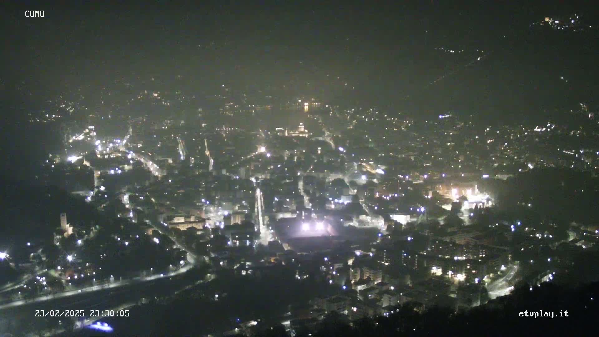 A nighttime, hazy view of a city illuminated by many lights, nestled between hills.