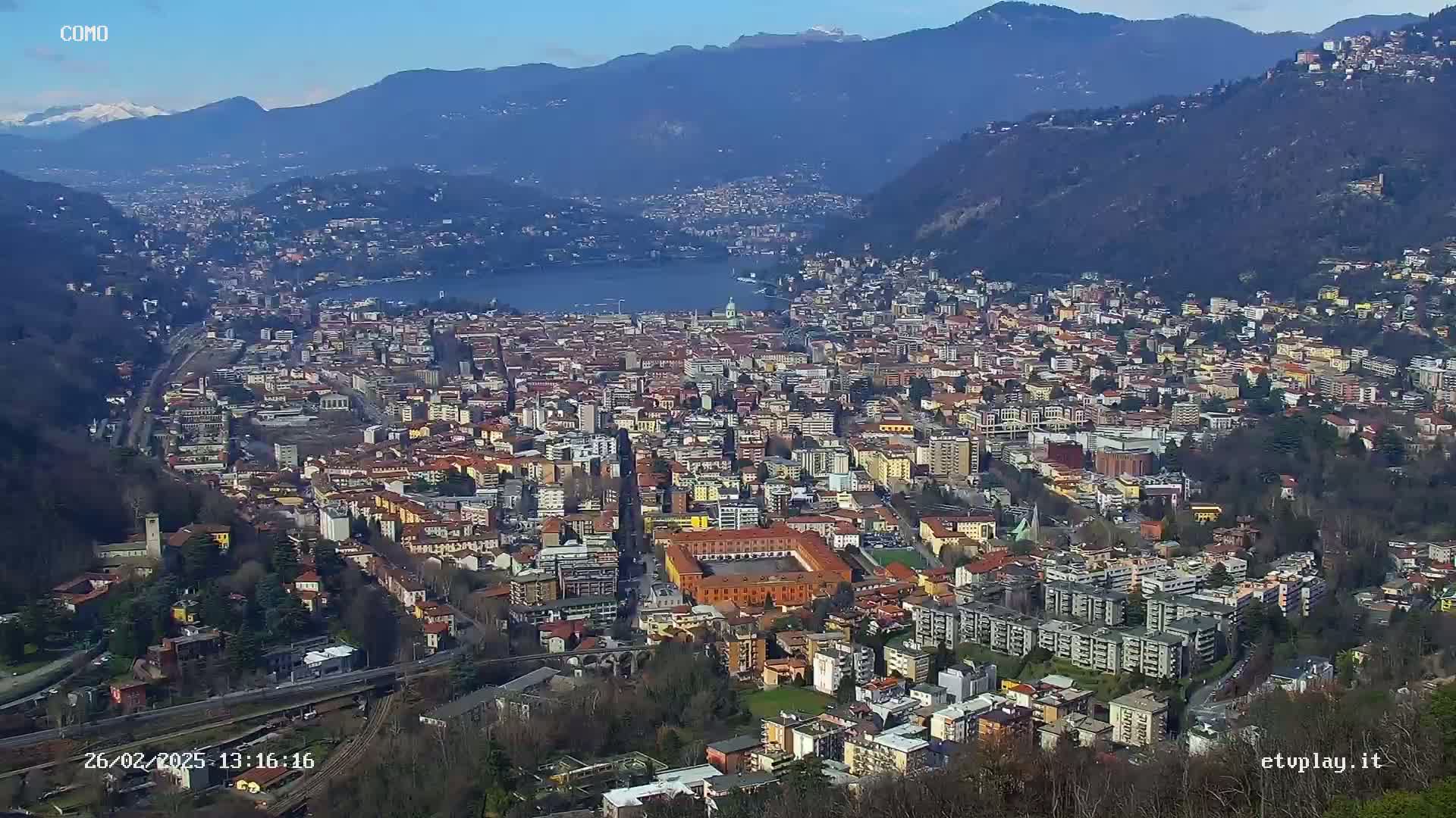 A sprawling city nestled between mountains and a lake, under a clear, sunny sky.
