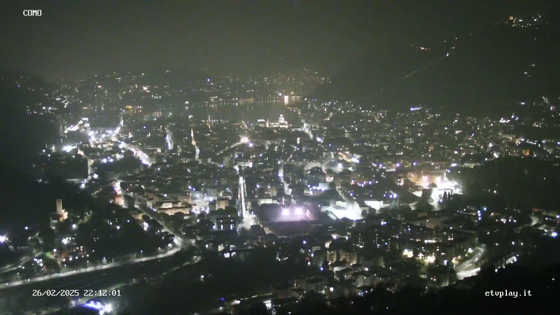 A nighttime, hazy aerial view of a city illuminated by numerous lights, situated beside a body of water.