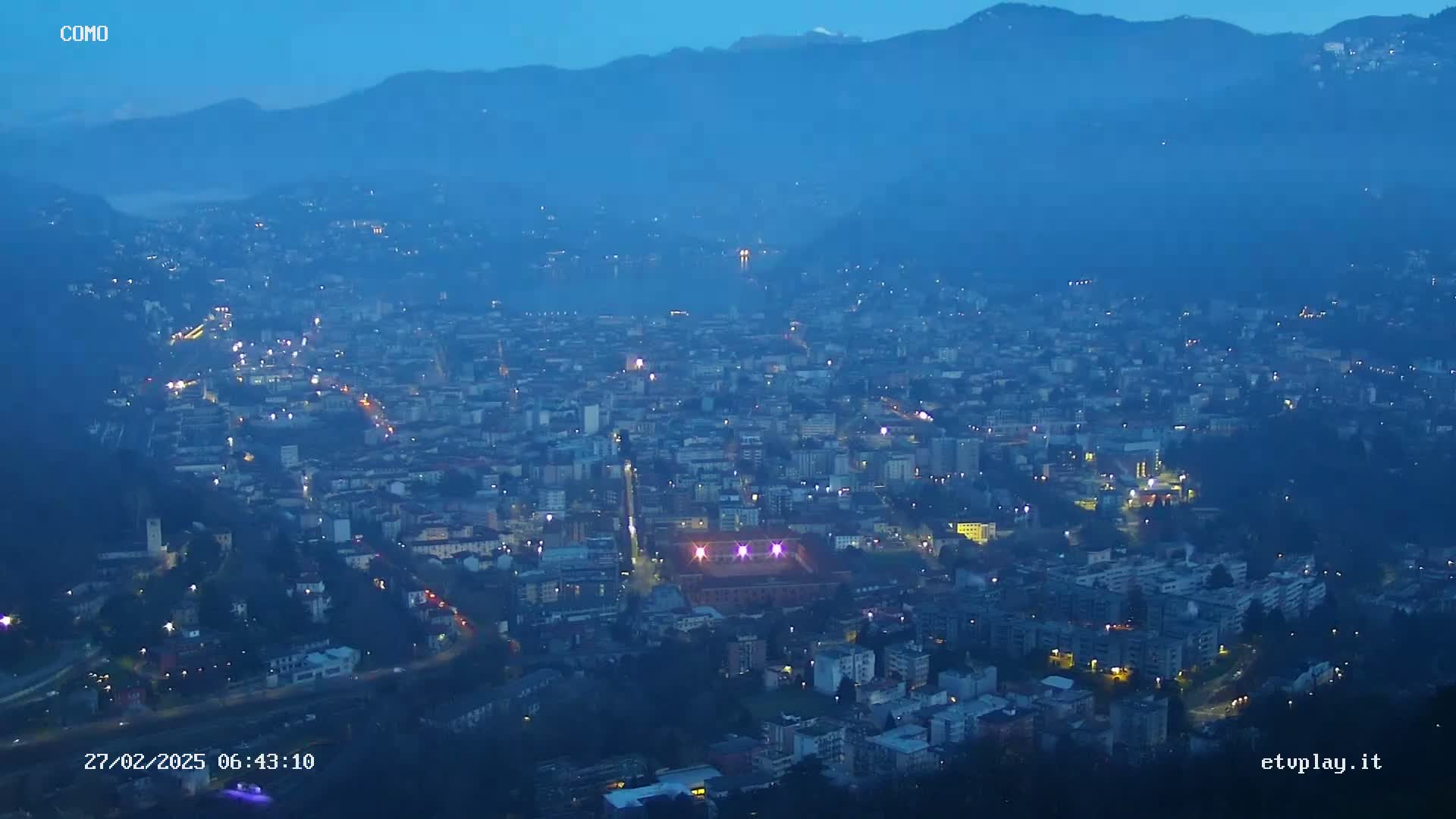 A sprawling city at twilight is nestled in a valley between mountains under a hazy, dark blue sky.