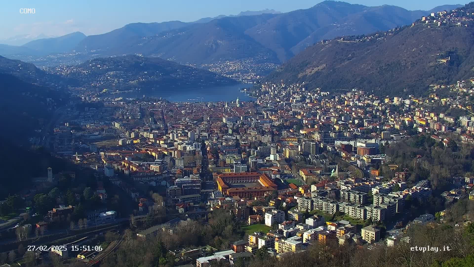 A sprawling city nestled between mountains and a lake under a clear, sunny sky.