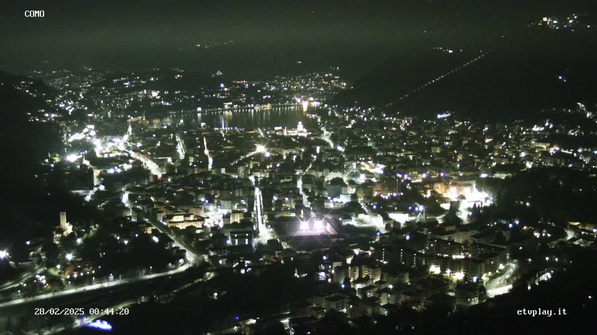 A nighttime, high-angle view of a city illuminated by streetlights, with a body of water visible in the distance under a hazy sky.