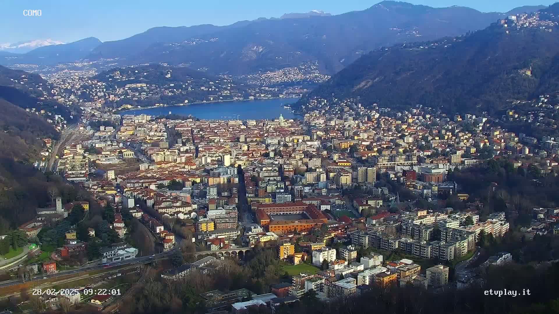 A sprawling city nestled between mountains and a lake under a clear, sunny sky.