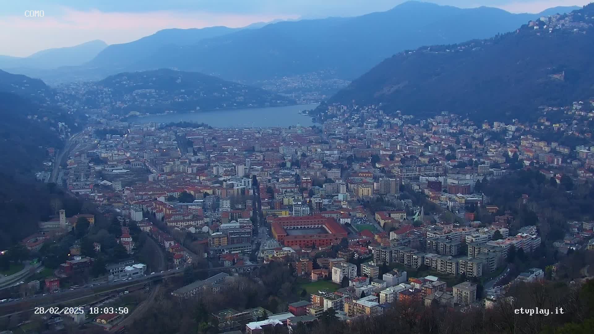 An expansive city nestled between a lake and mountains under a twilight sky.