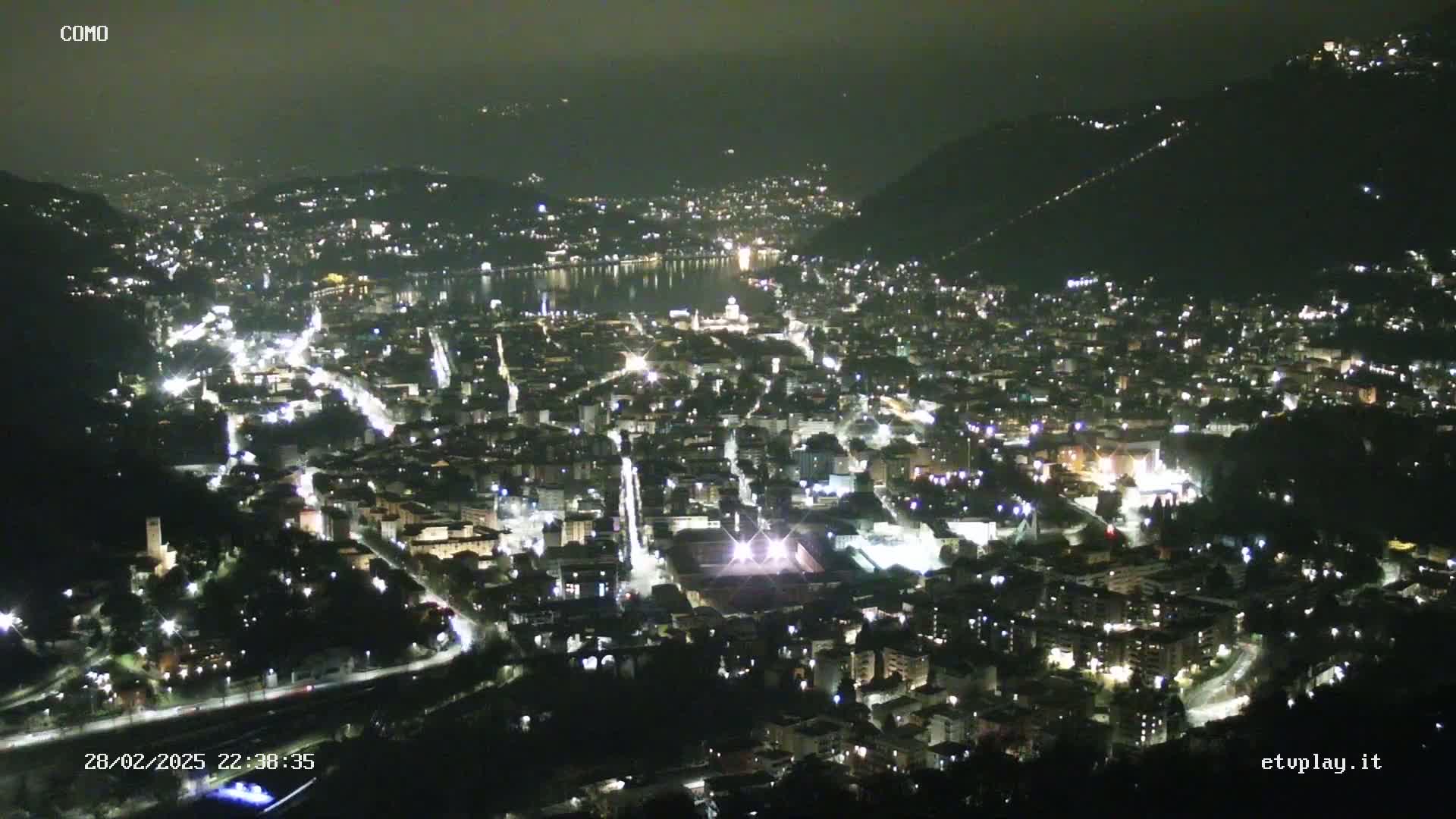 A nighttime, high-angle view of a city nestled between hills and a body of water, illuminated by numerous lights.