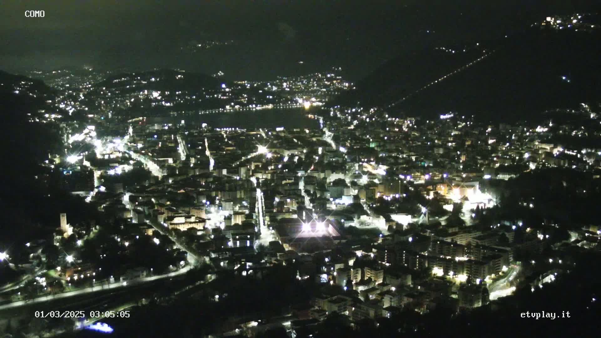 A nighttime aerial view of a city illuminated by streetlights, nestled in a valley with a dark body of water visible in the distance.