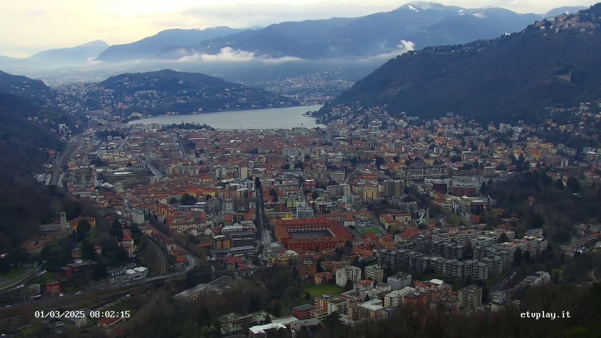 A sprawling city nestled between mountains and a lake under a cloudy sky.