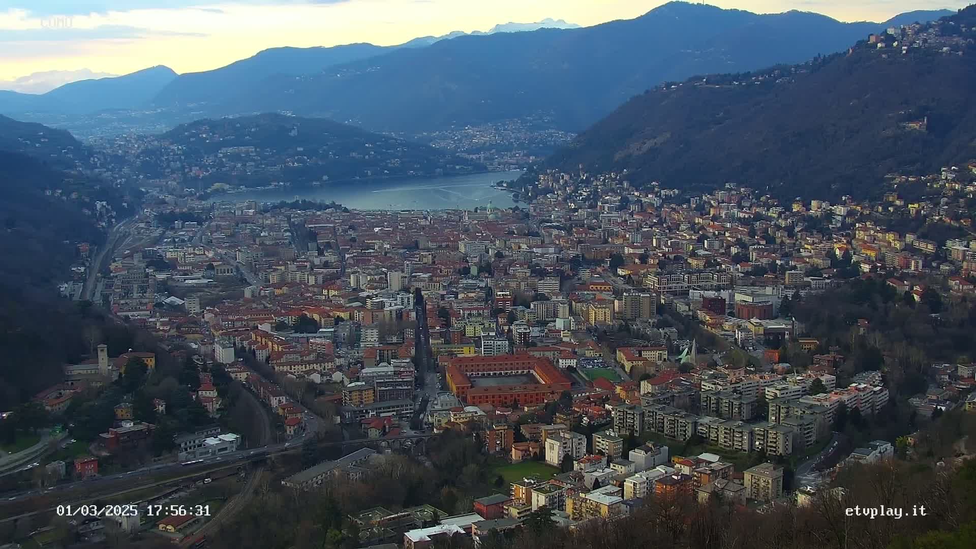 A sprawling city nestled in a valley between mountains, overlooking a lake under a partly cloudy sky.