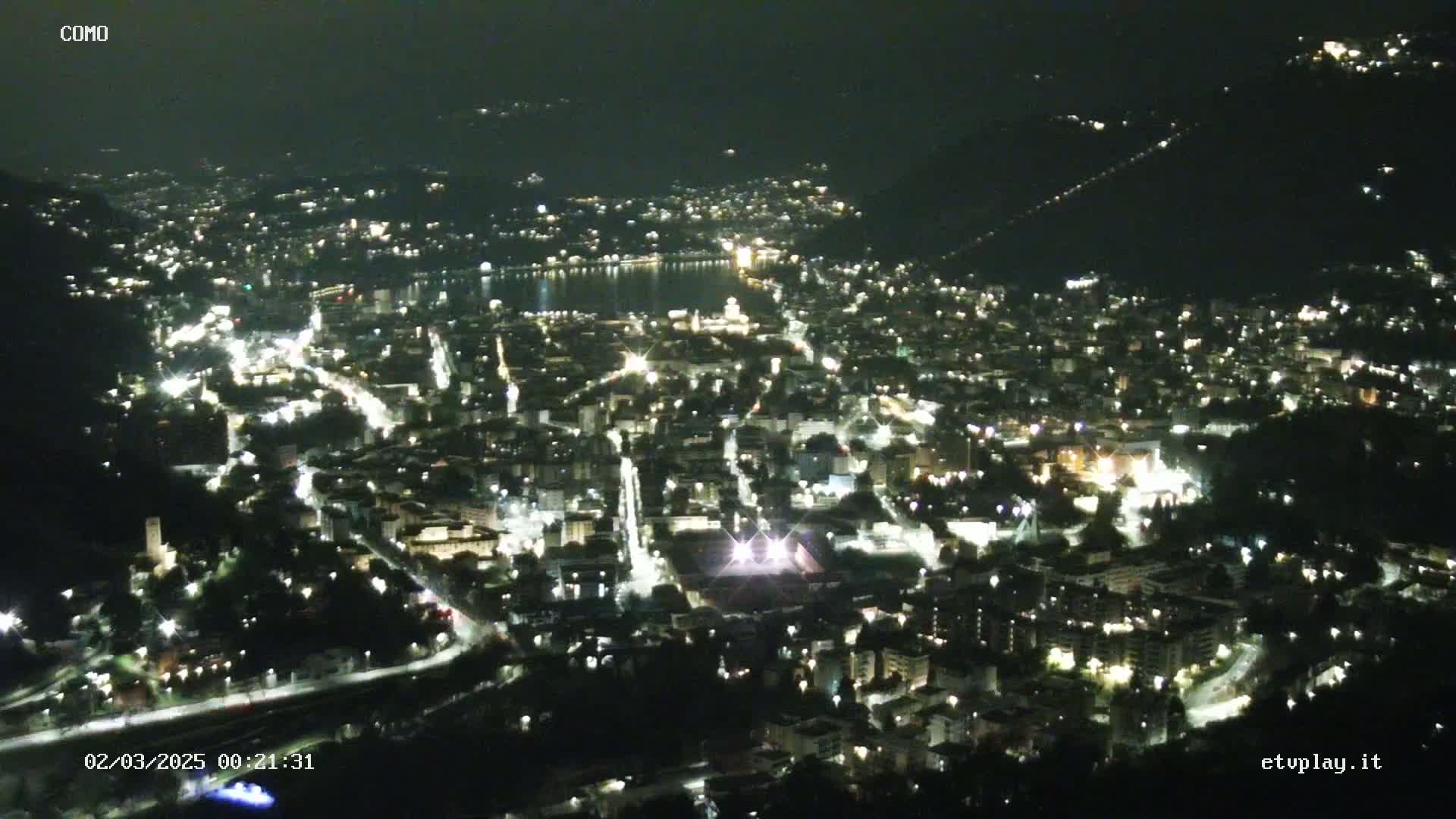 A nighttime, aerial view of a city nestled beside a dark body of water, illuminated by numerous lights.