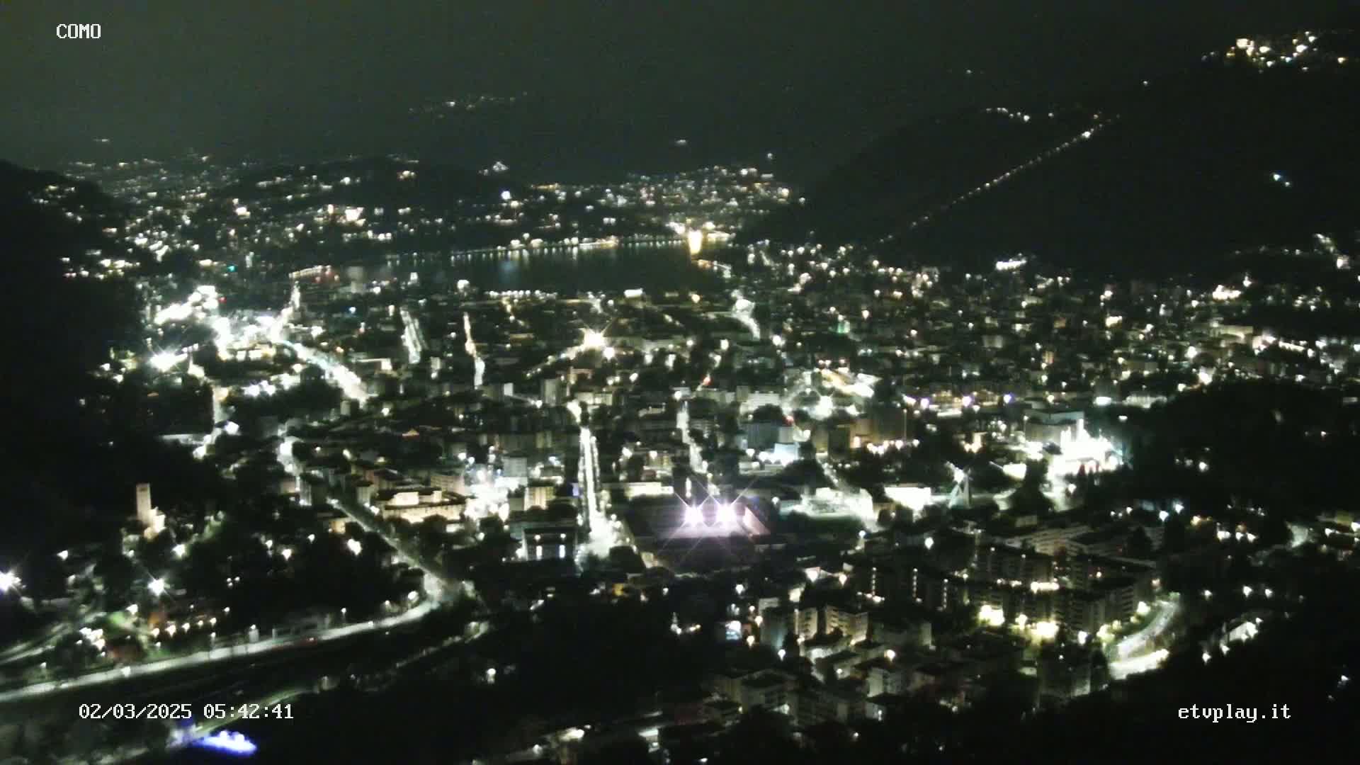 A nighttime aerial view shows a city illuminated by lights, nestled beside a dark body of water and surrounded by hills under a clear, dark sky.