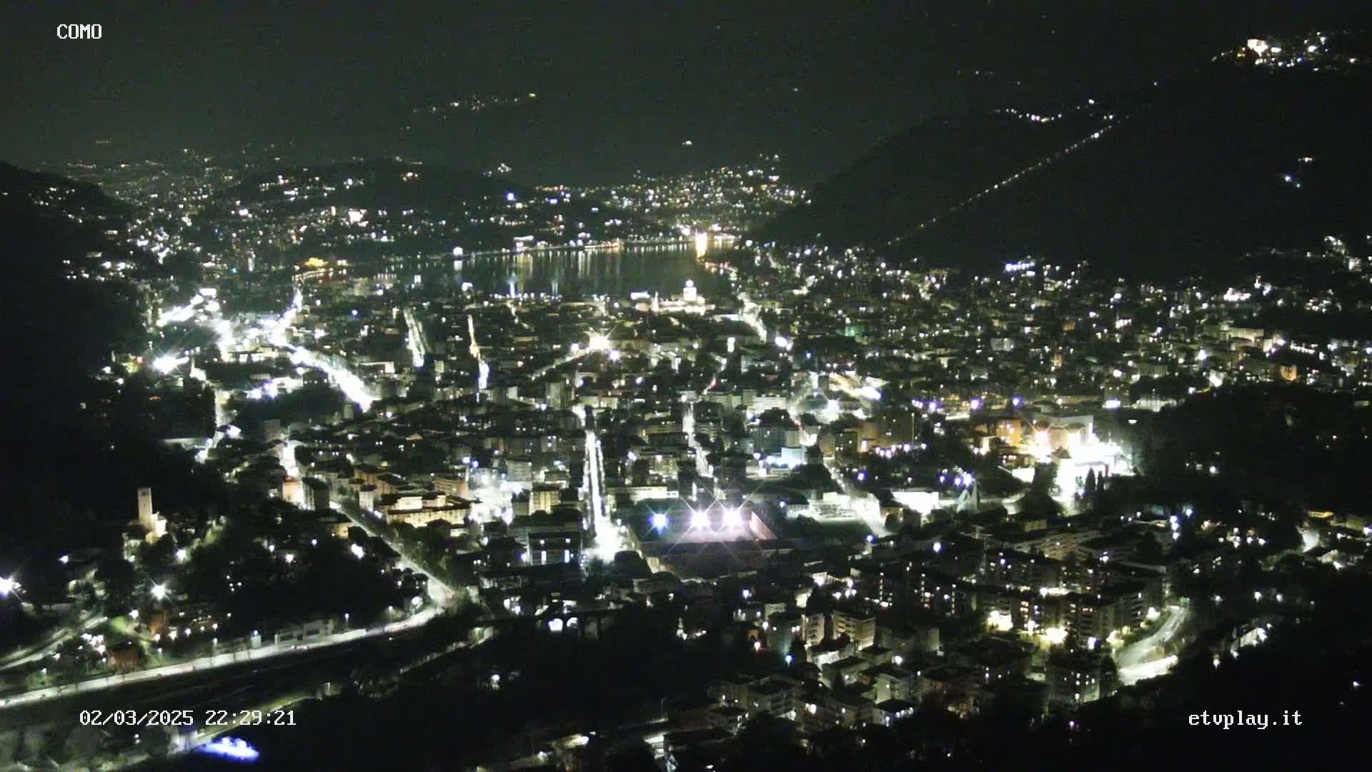 A nighttime aerial view of a city nestled beside a body of water, illuminated by streetlights and building lights.