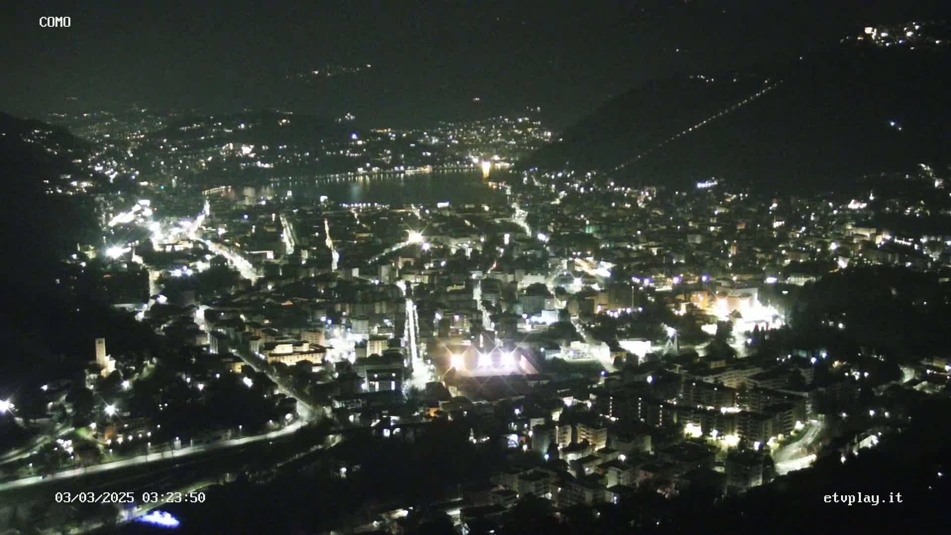 A nighttime, aerial view of a city nestled beside a lake, illuminated by numerous streetlights.