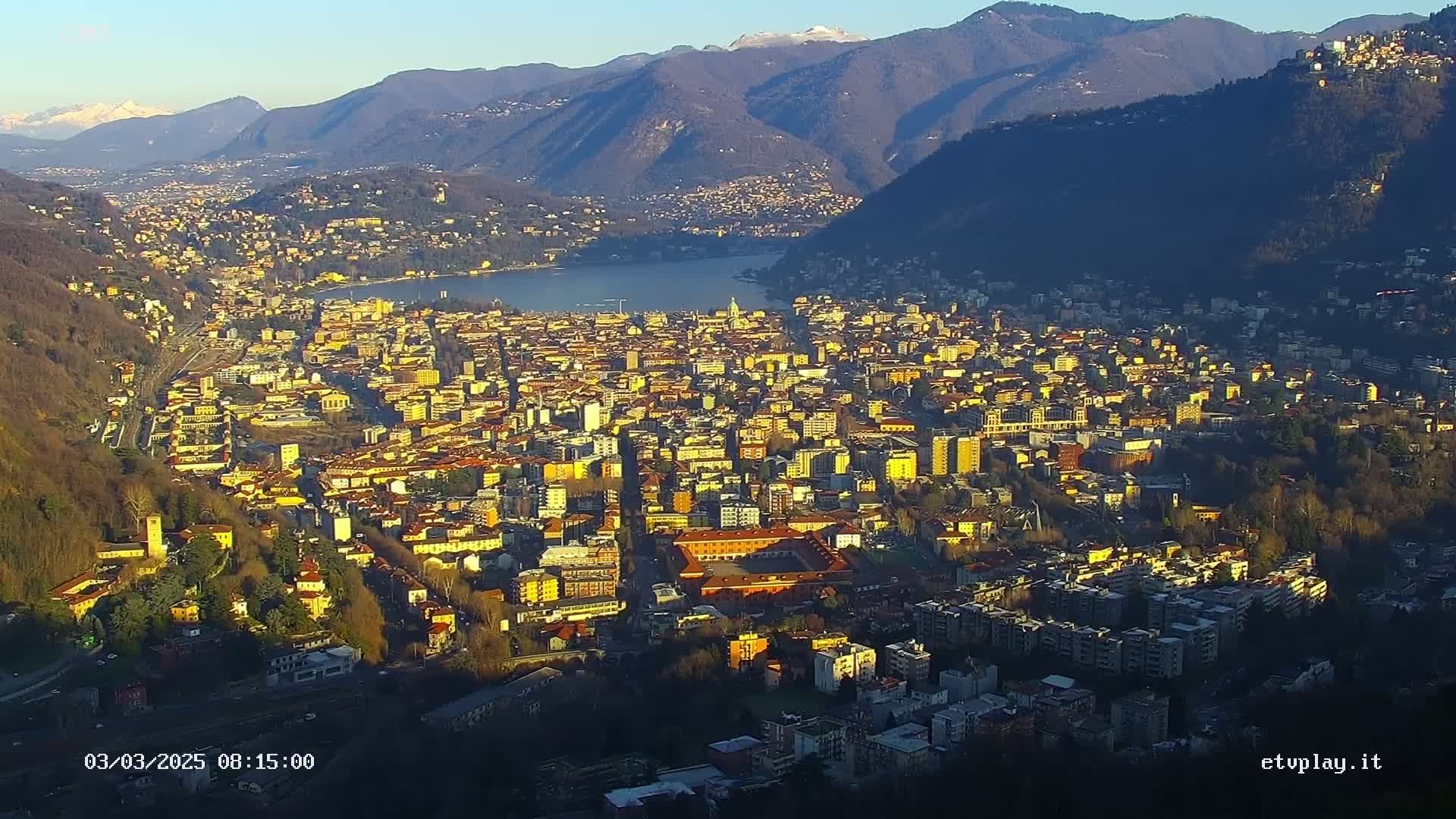 A sprawling city nestled between mountains and a lake, bathed in the golden light of a clear, sunny day.