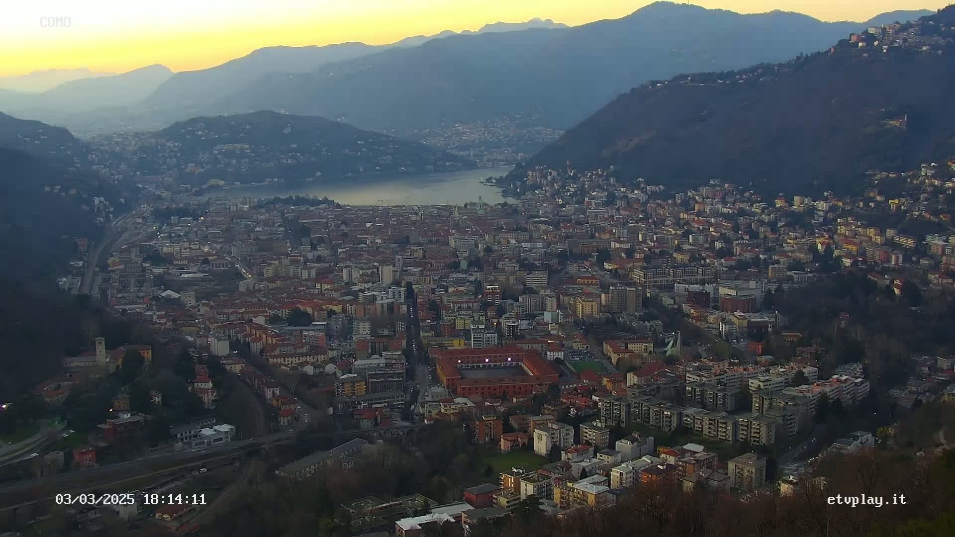 A sprawling city nestled between mountains and a lake, viewed at sunset under clear skies.