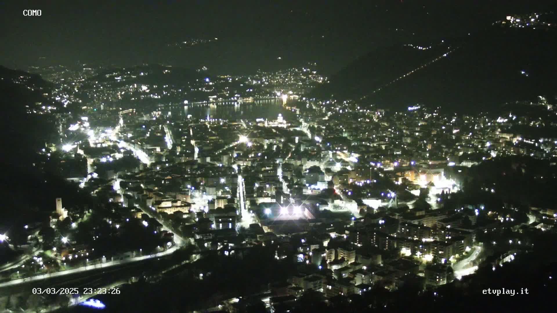 A nighttime, high-angle view shows a city illuminated by streetlights and building lights, nestled beside a dark body of water under a clear, dark sky.