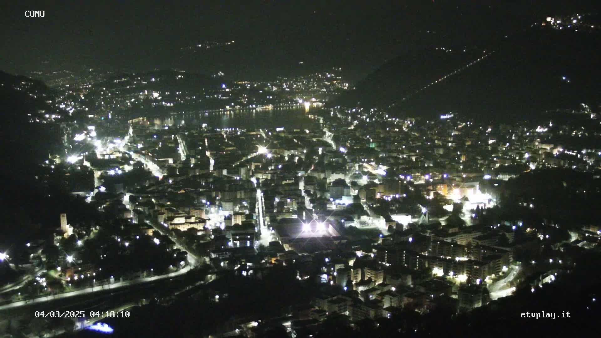 A nighttime, high-angle view of a city illuminated by streetlights and building lights, nestled beside a dark body of water under a clear, dark sky.