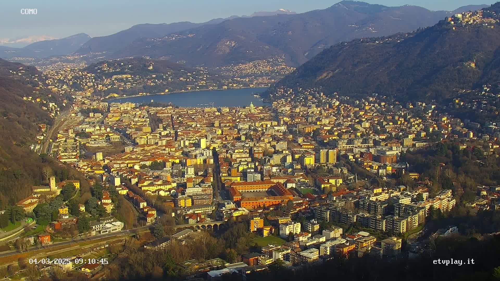 A sprawling city nestled beside a lake, surrounded by mountains under a clear, sunny sky.