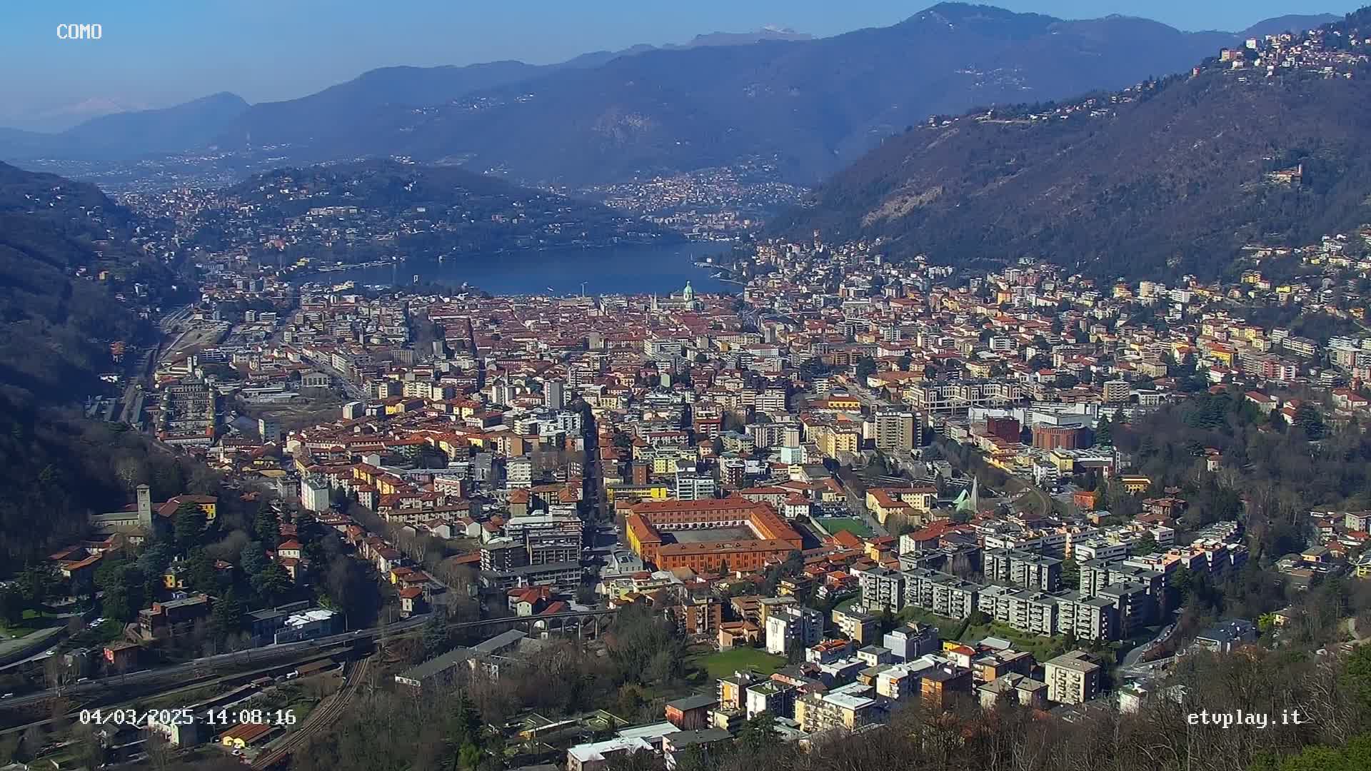 A sprawling city nestled in a valley between mountains, overlooking a lake under a clear, sunny sky.