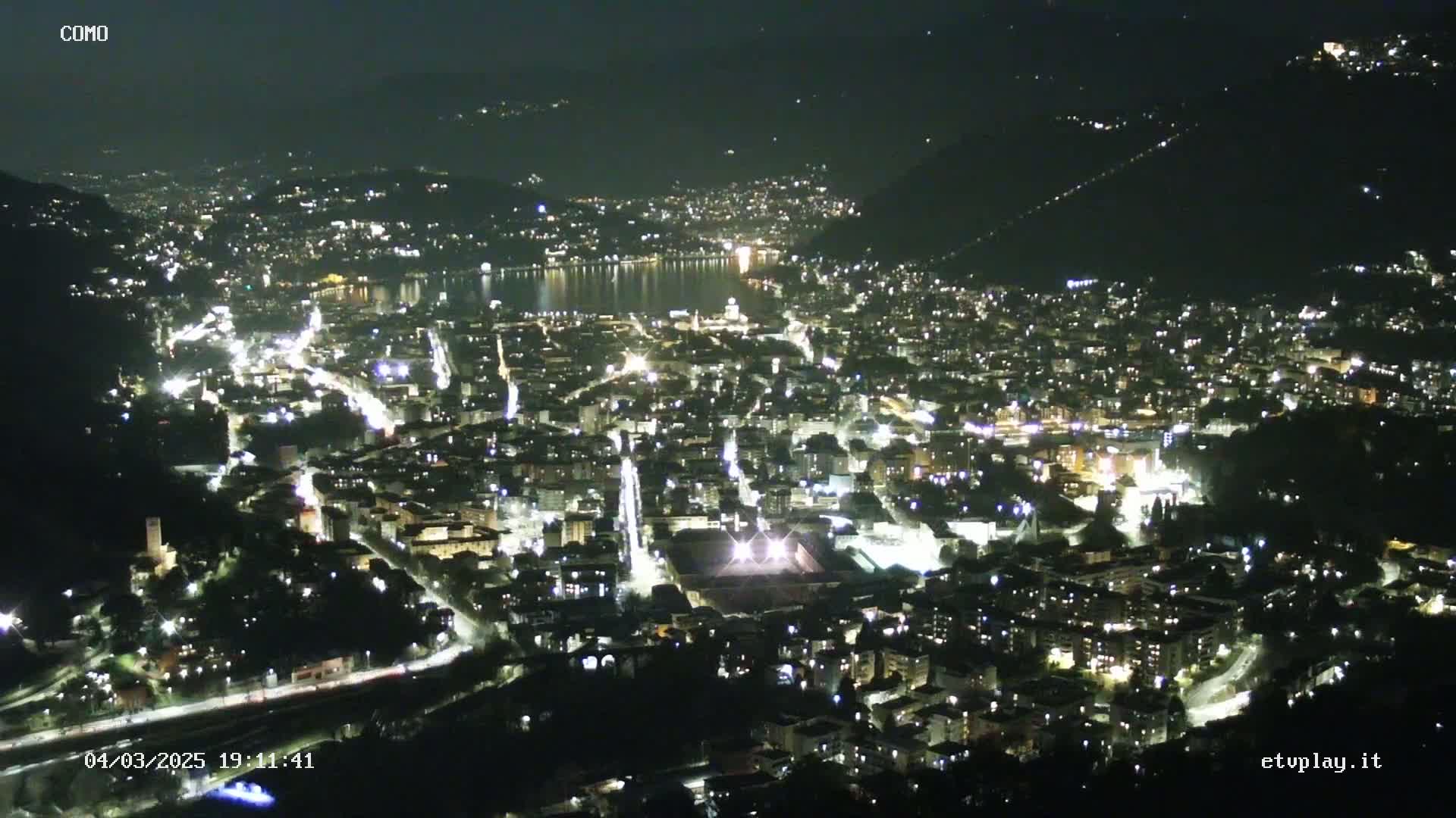 A nighttime aerial view of a city nestled beside a body of water, illuminated by streetlights and building lights.