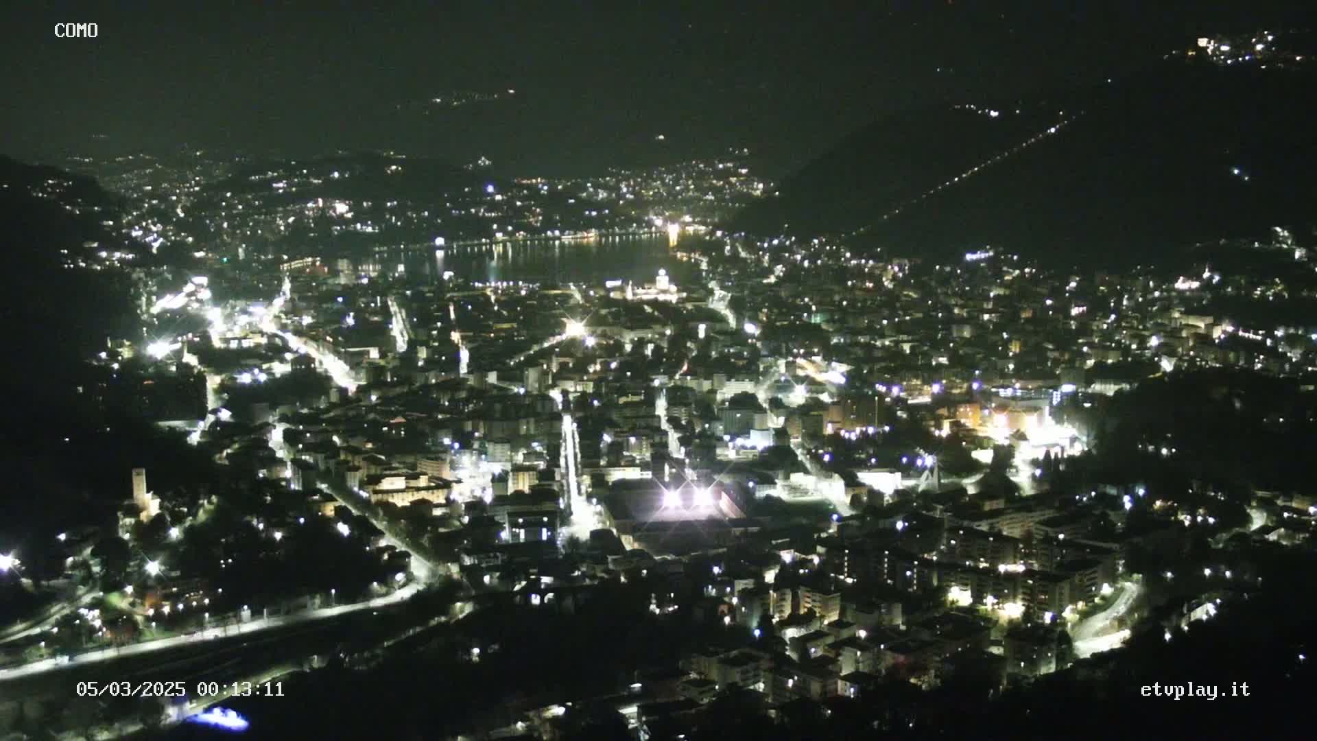 A nighttime aerial view of a city illuminated by streetlights, nestled between hills and a body of water under a clear, dark sky.