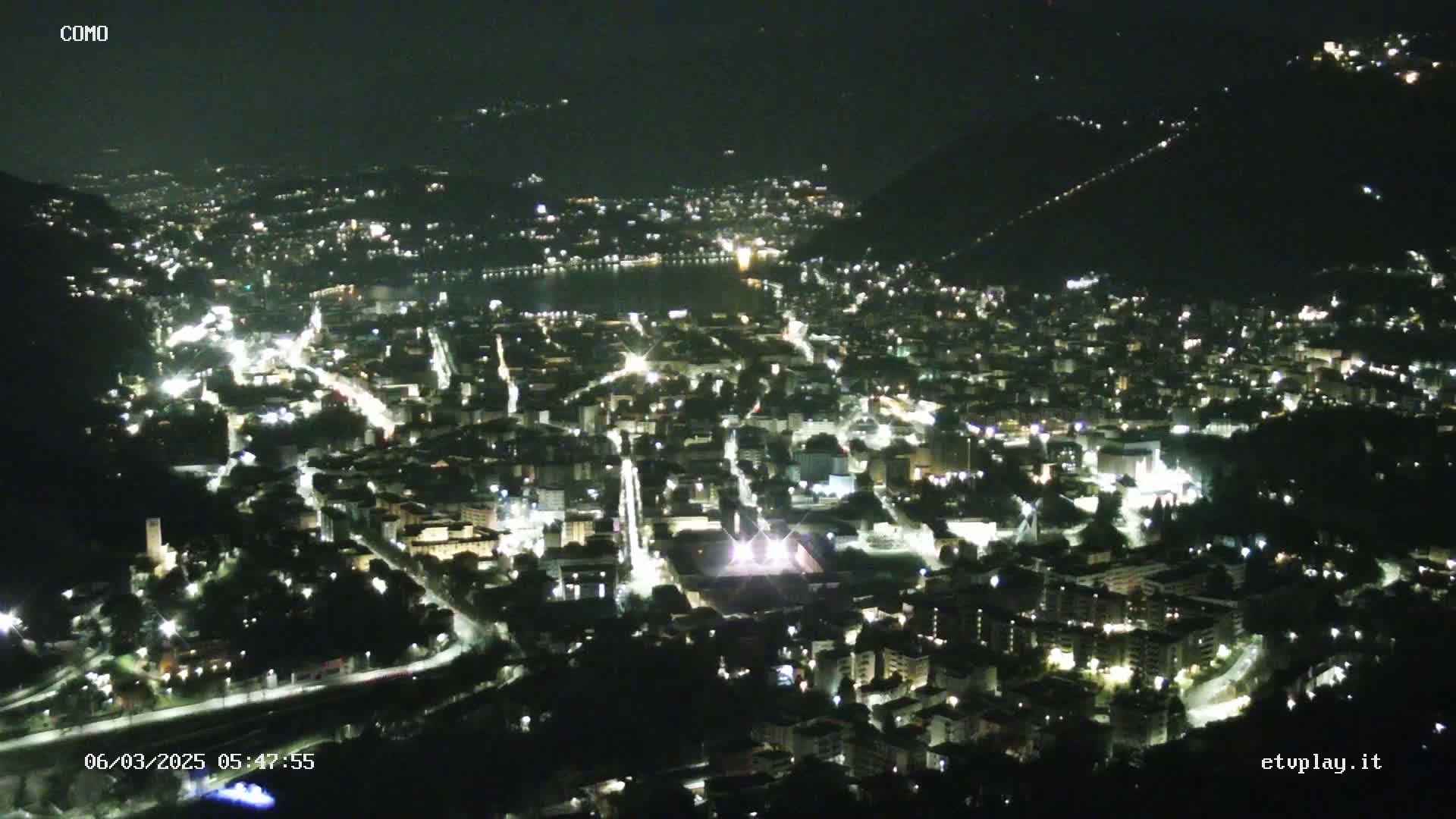 A nighttime, high-angle view of a city nestled in a valley, illuminated by numerous streetlights, with a body of water visible in the distance.