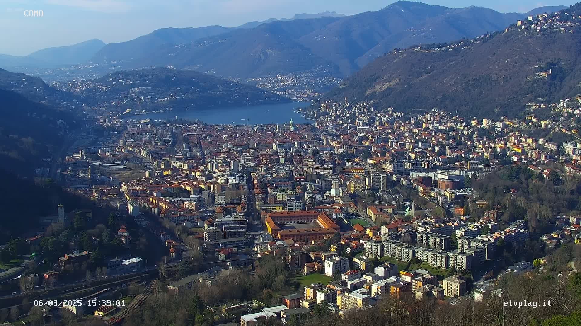 A sprawling city nestled between mountains and a lake under a clear, sunny sky.