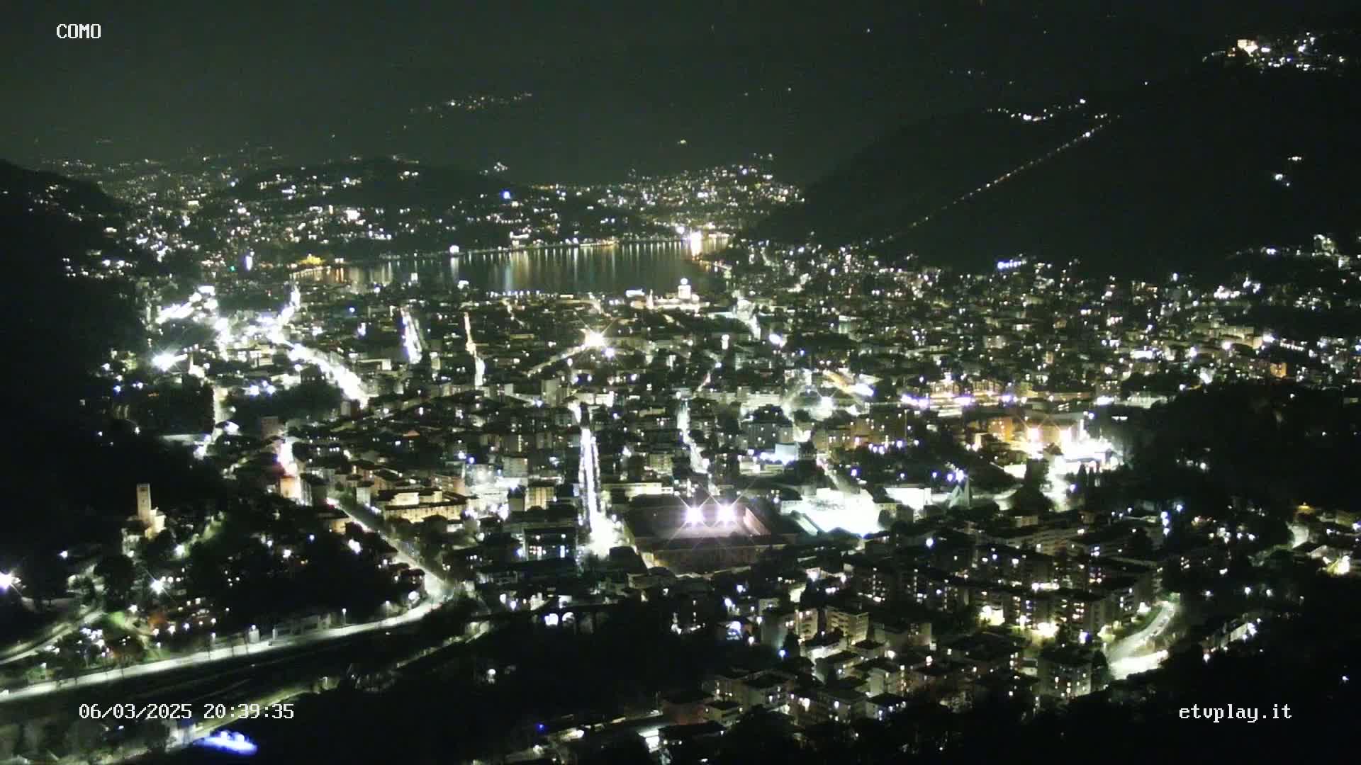 A nighttime aerial view of a city illuminated by streetlights, nestled beside a dark body of water under a clear, dark sky.