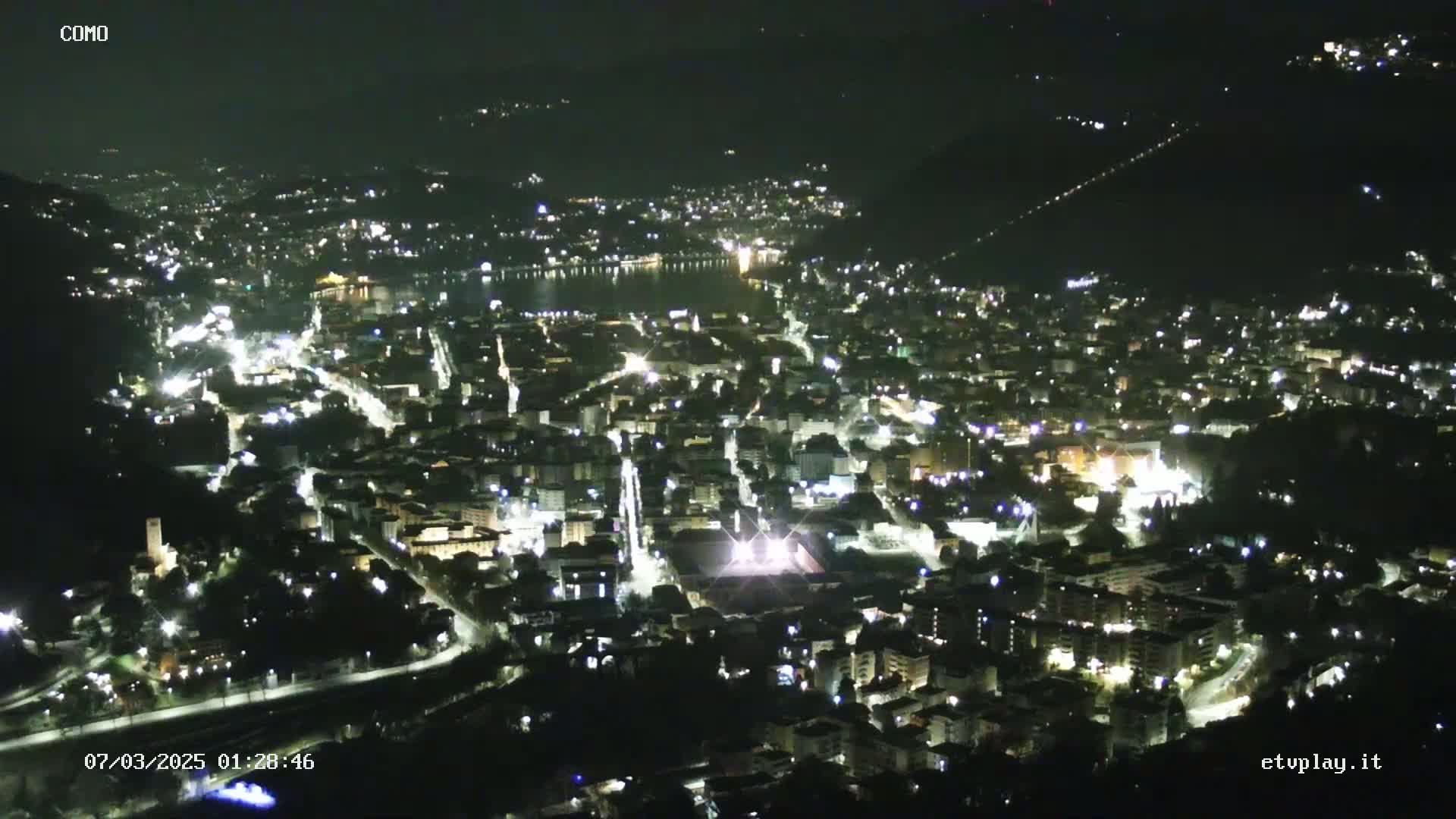 A nighttime, high-angle view of a city illuminated by streetlights and building lights, situated near a body of water.