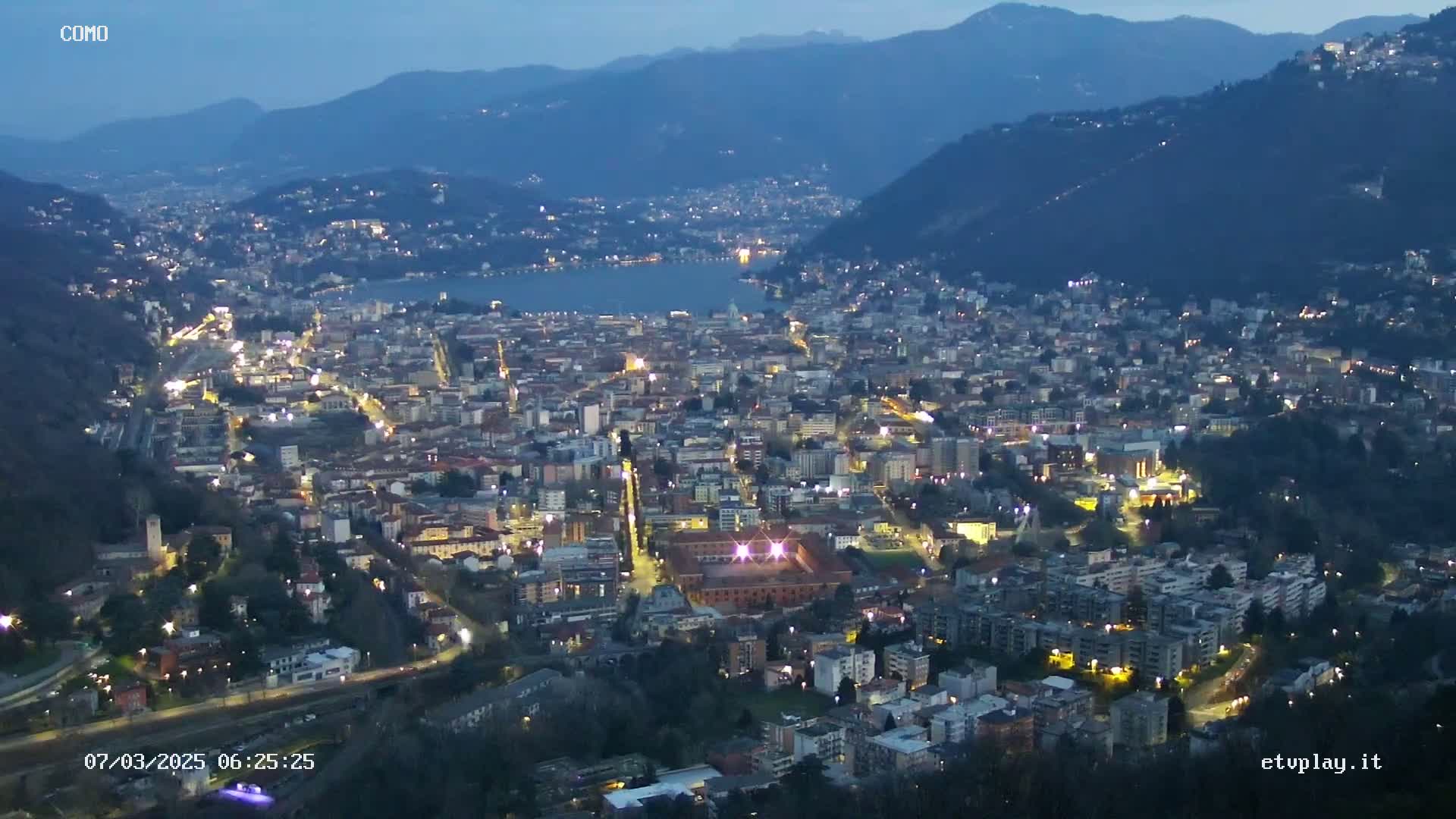 A nighttime aerial view of a city nestled in a valley beside a lake, under a twilight sky.