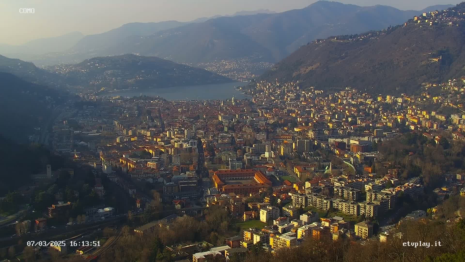 A hazy, sunny day reveals a sprawling city nestled in a valley between mountains, with a lake visible in the distance.