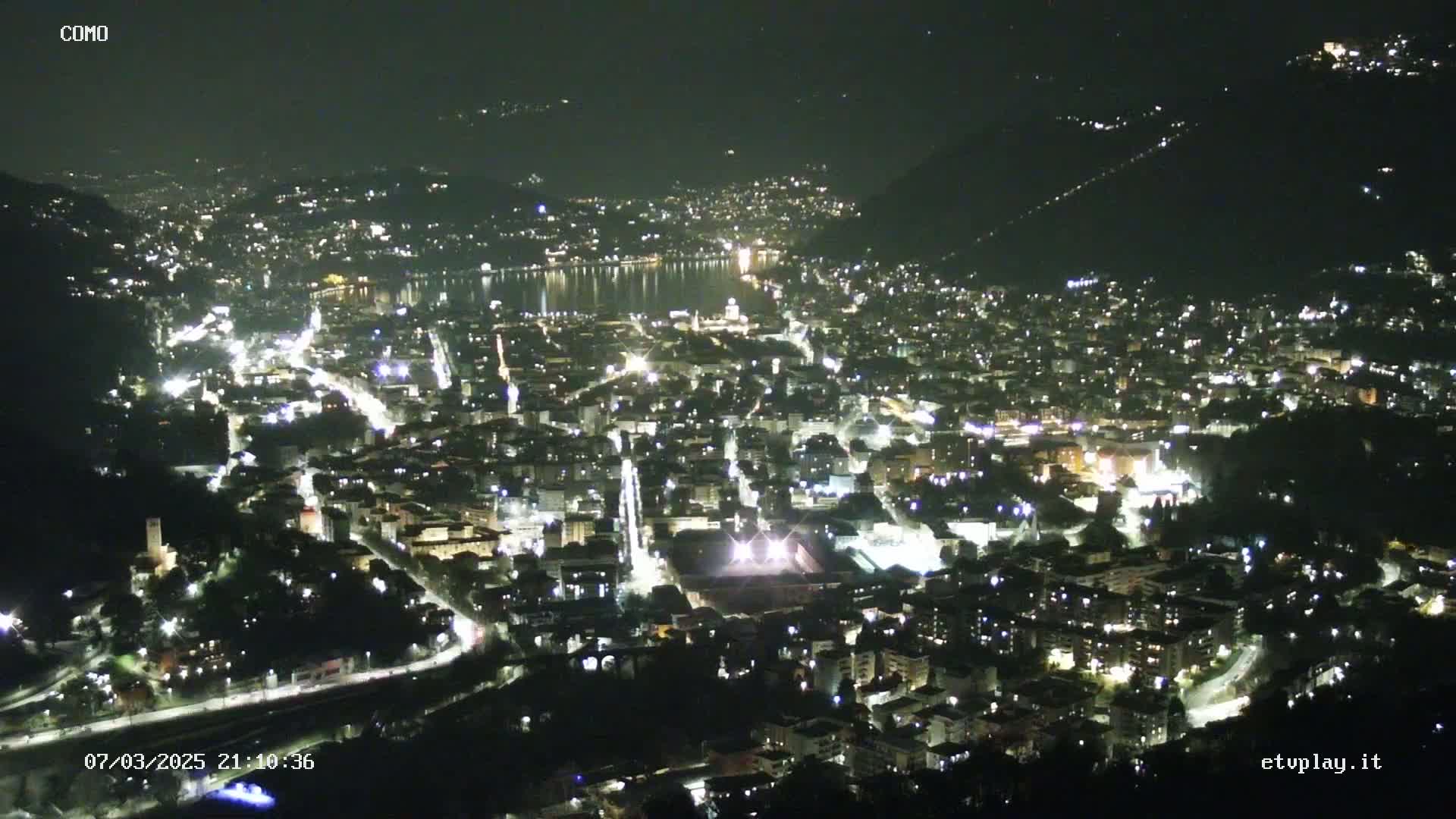 A nighttime aerial view of a city illuminated by lights, situated near a body of water under a clear sky.
