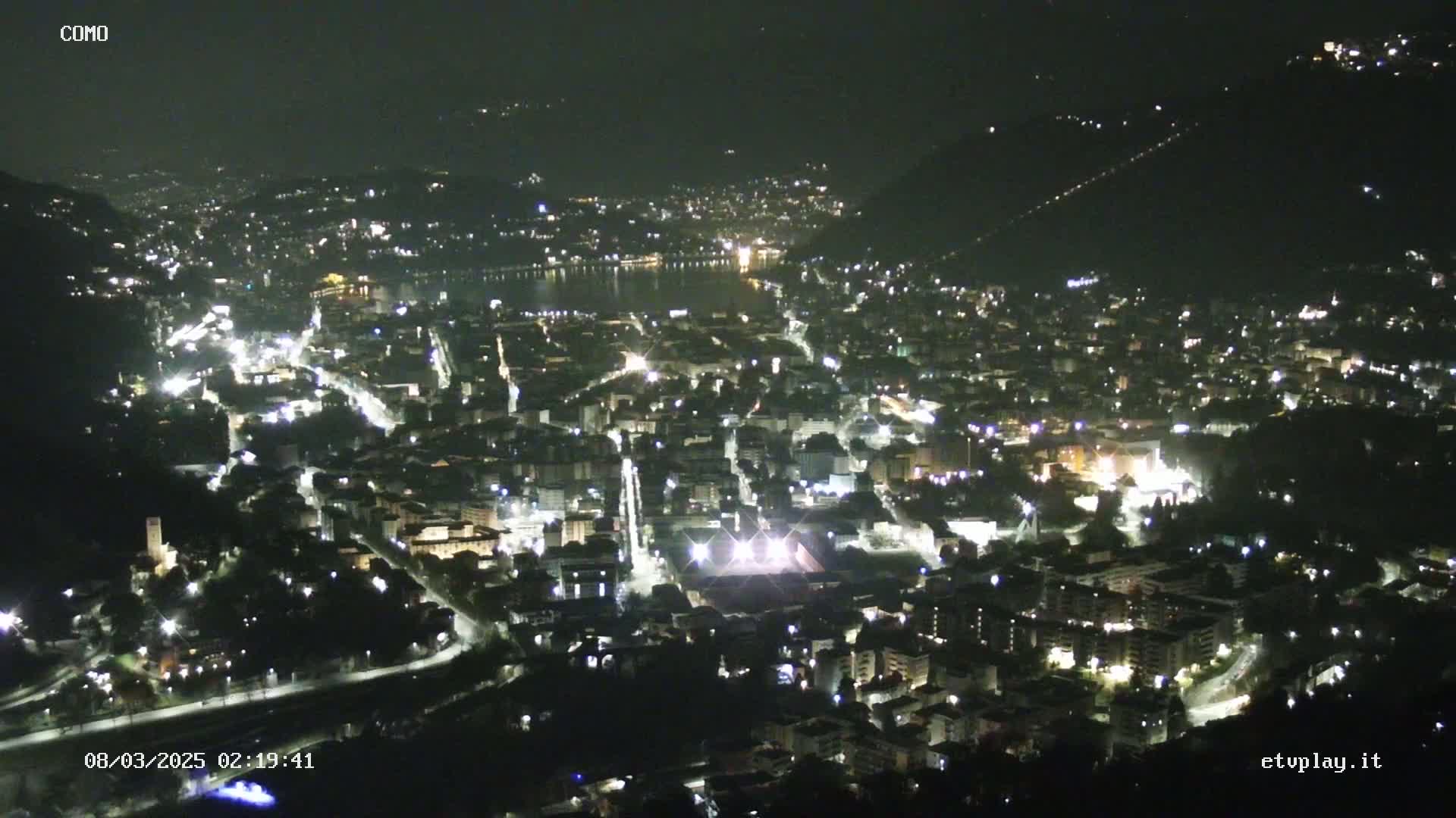 A nighttime aerial view of a city nestled between hills, with a body of water visible in the distance under a clear sky.