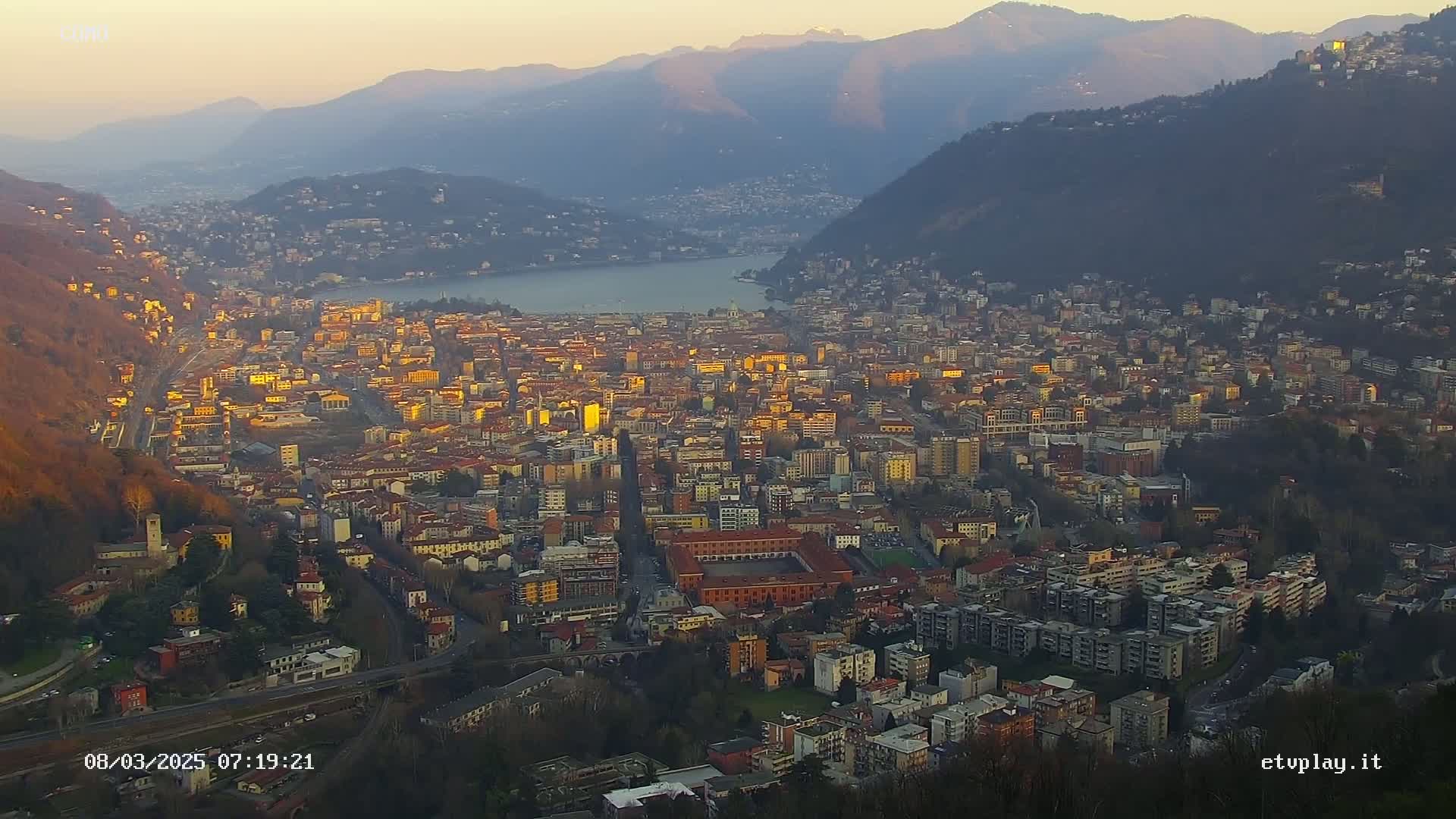 A sprawling city nestled in a valley between mountains, bathed in the golden light of a setting sun, overlooks a calm lake.