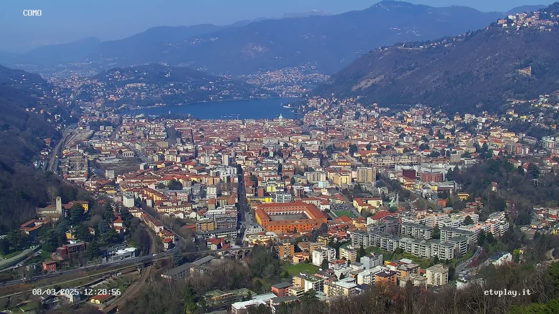 A sprawling city nestled in a valley between mountains, with a lake visible in the distance, under a clear, sunny sky.